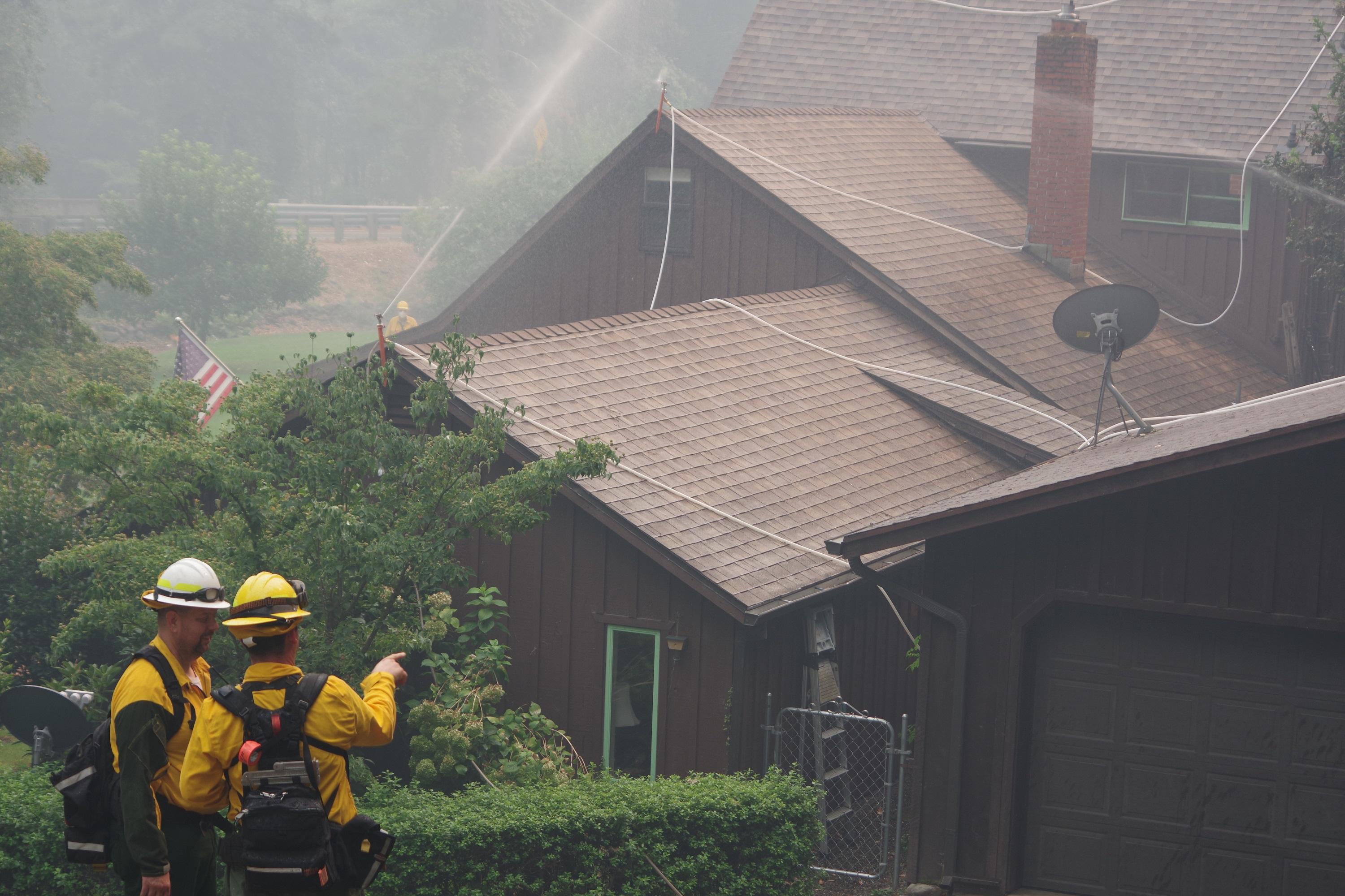 A series of sprinklers are spraying water on the roof and sides of a large house. Two firefighters in the foreground are looking to see if the sprinklers are working well.