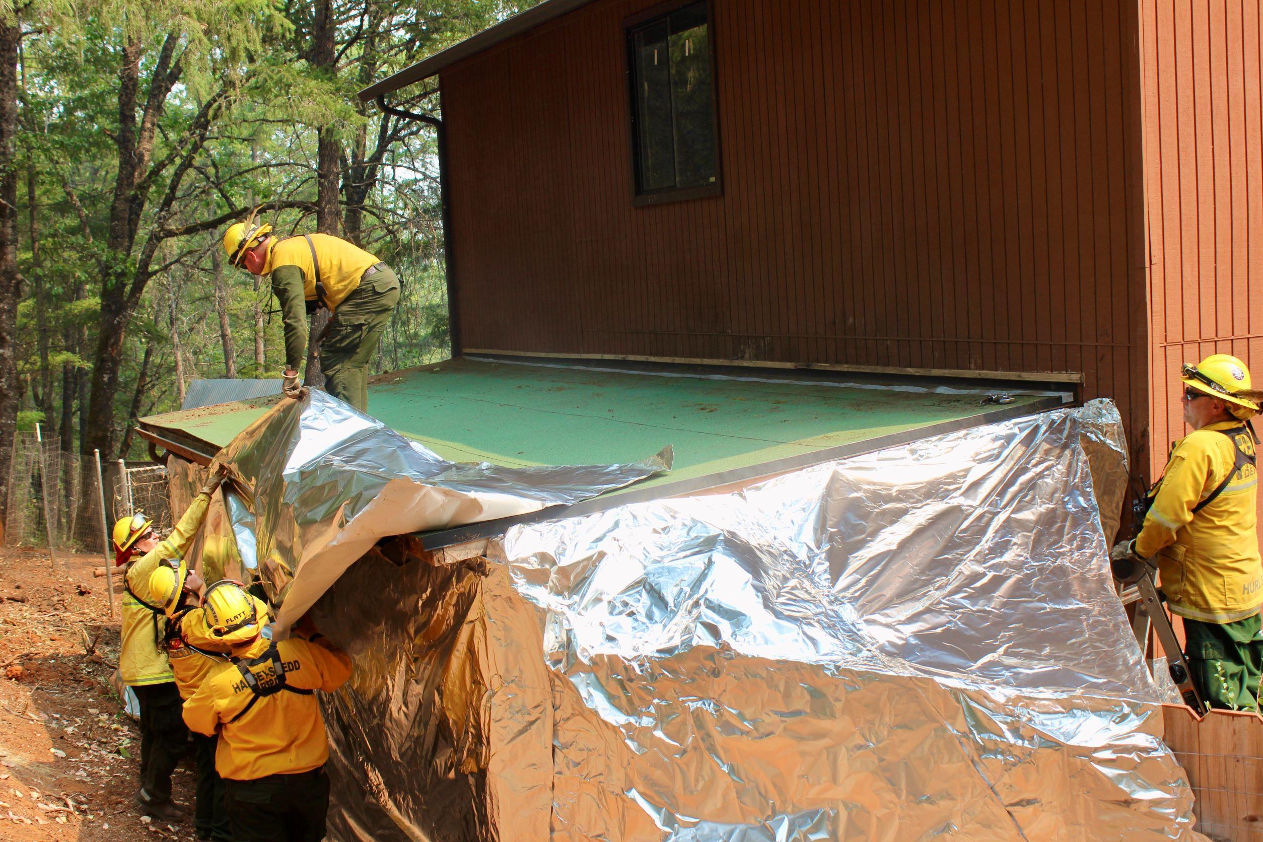 Four firefighters are wrestling structure wrap into place over the back of a structure. A fifth firefighter watches from on a ladder to the side. The structure is against the edge of the forest.