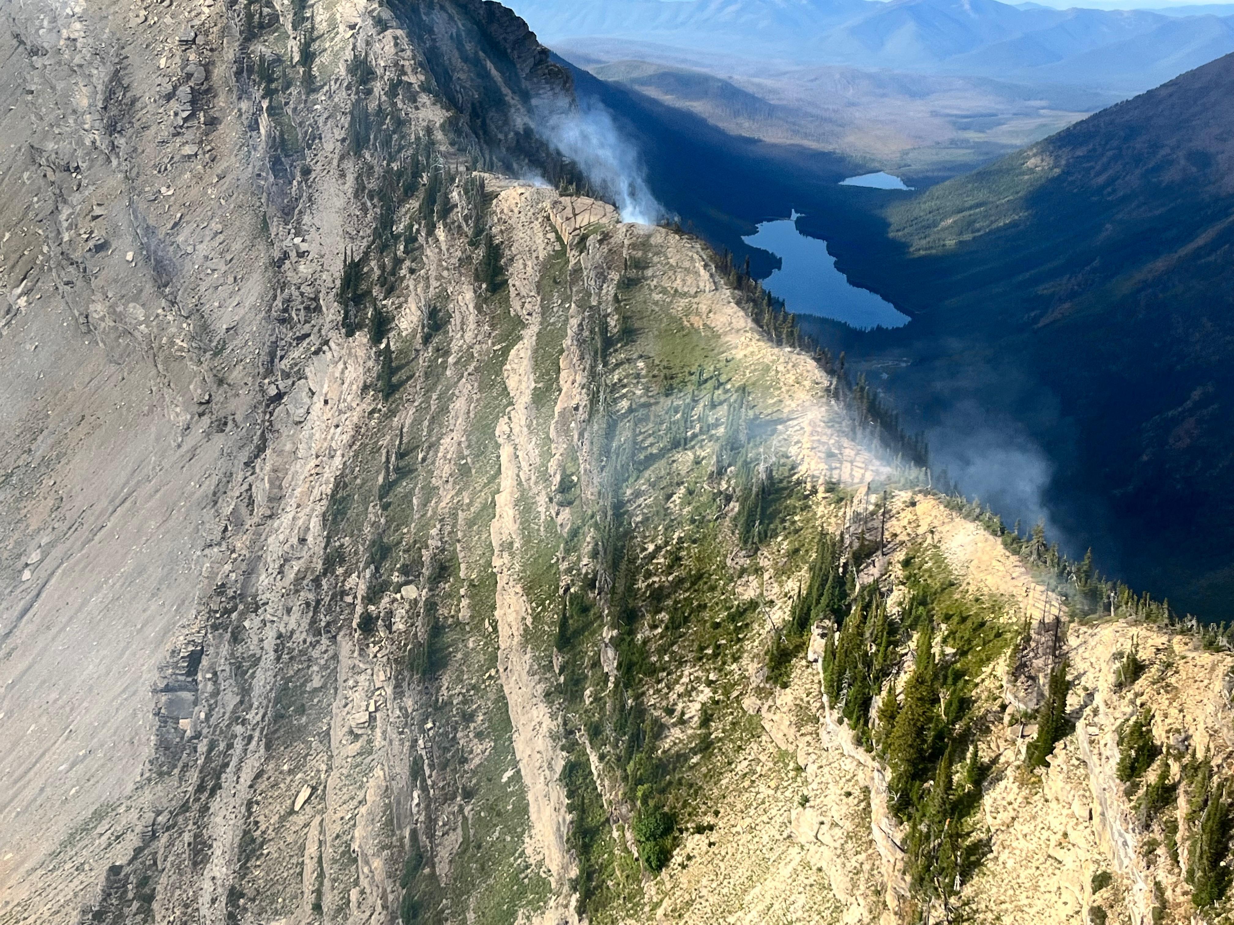 Aerial view of a high elevation rugged peak with small wisps of smoke coming from the ridge. Two lakes visible in the distance.