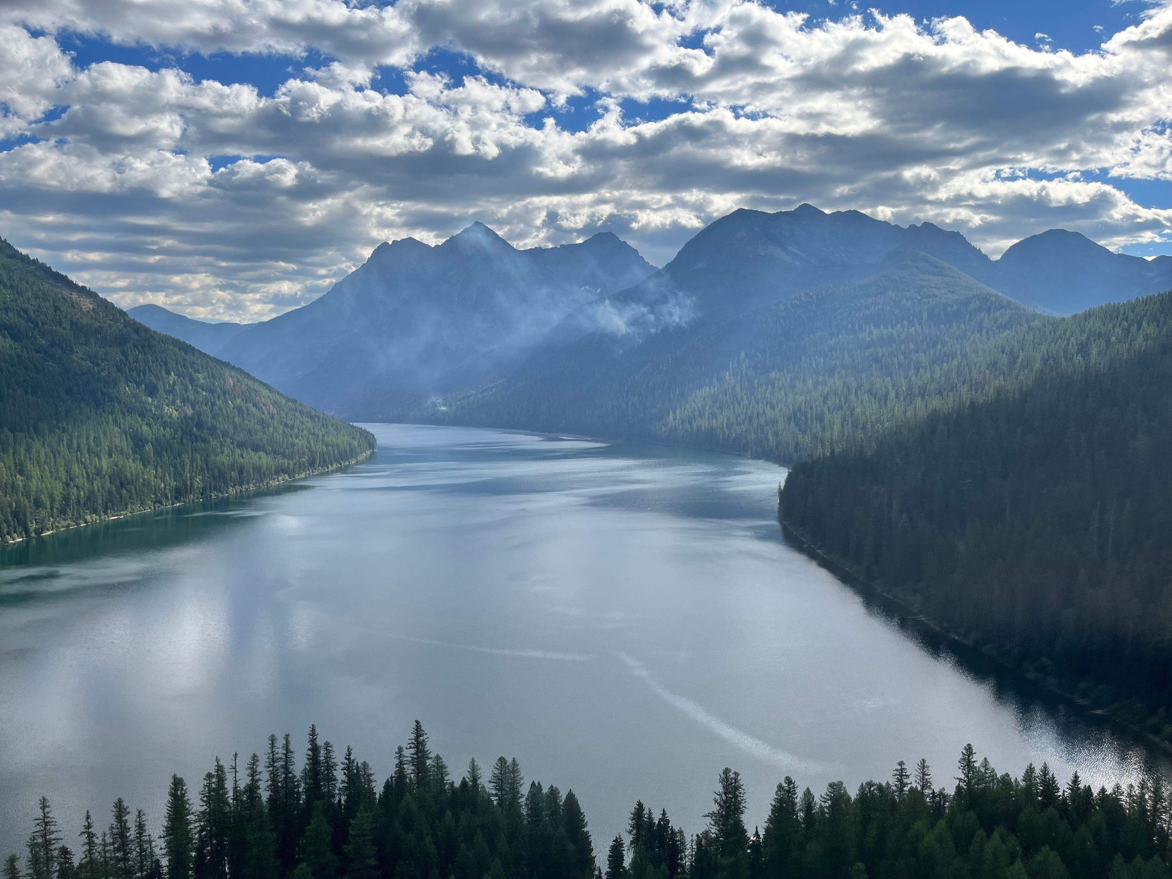 A long lake surrounded by mountains with white wisps of smoke coming from the forest on the right side of the lake