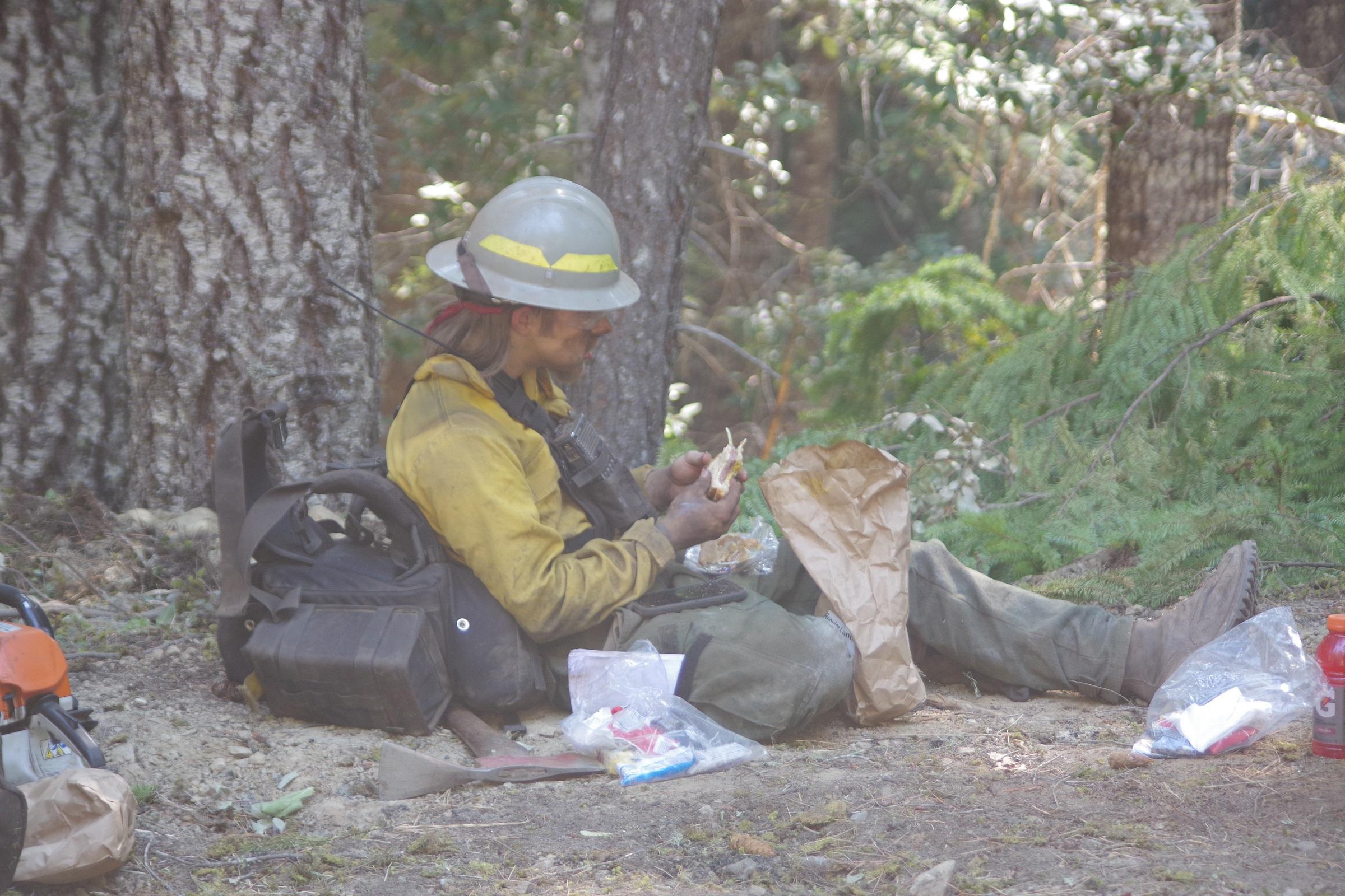 A firefighter is sitting on the ground in the dirt and ashes, eating a sandwich.