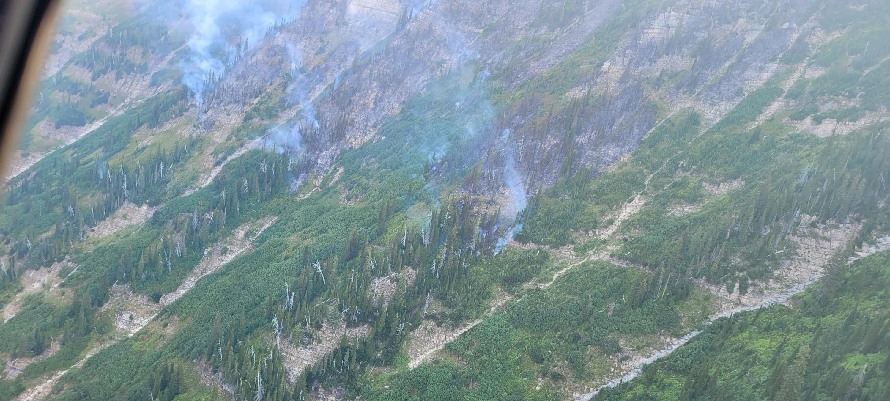 A photo shot from a helicopter showing smoke rising from fire smoldering in sparse trees interspersed with shrubs.