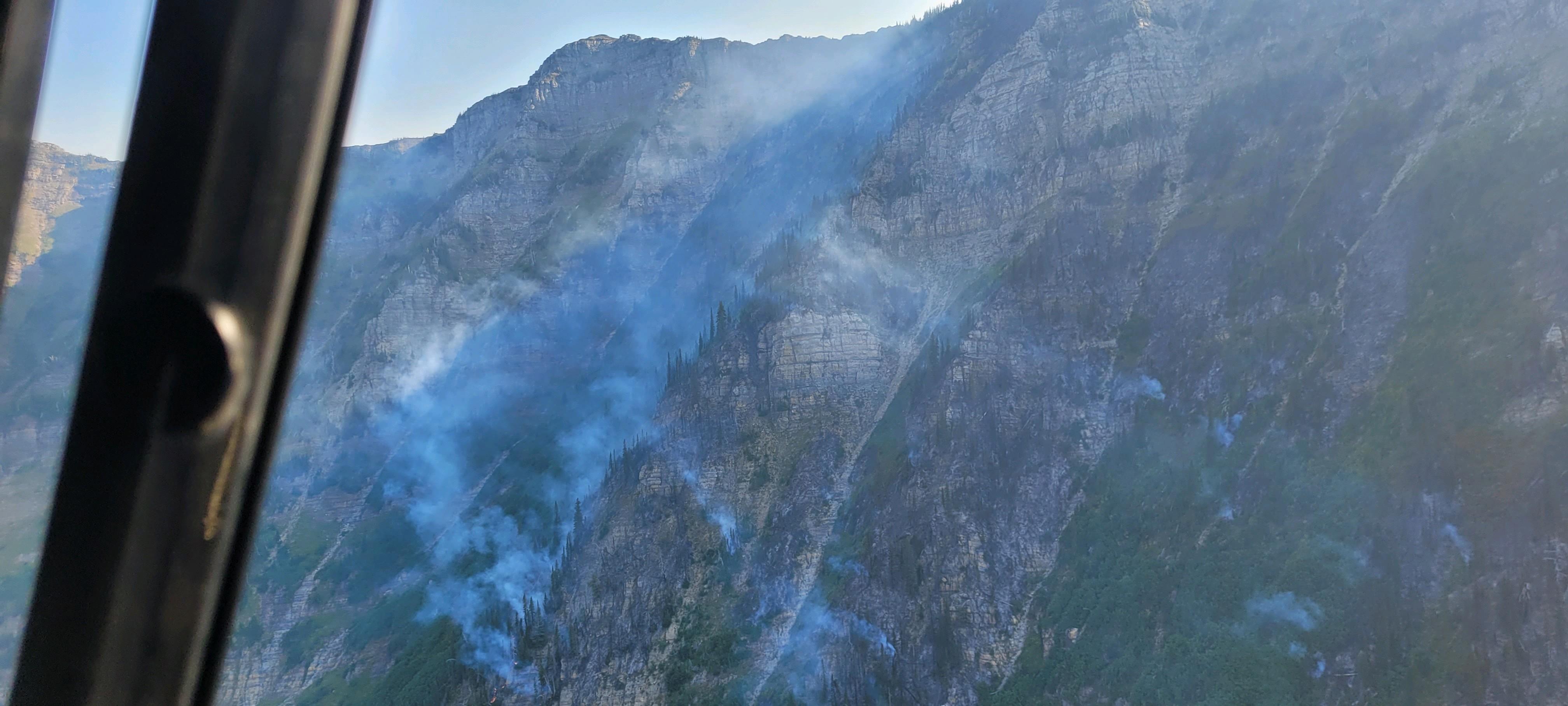 A photo shot from a helicopter showing steep slopes with smoke rising from several spots, with flames barely visible in one area.