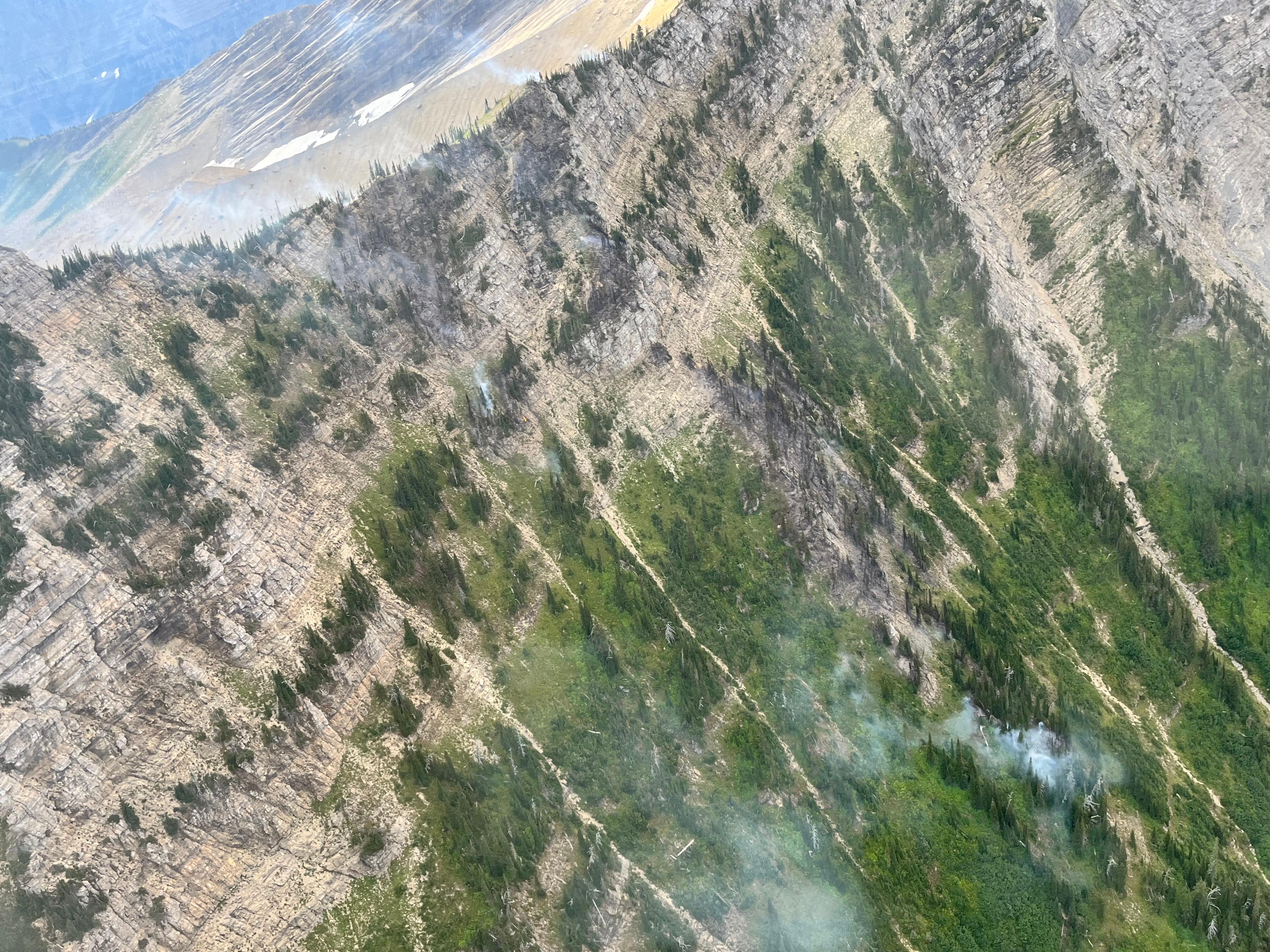 A photo of steep, rocky slopes with more vegetation below, where smoke rises from multiple clumps of trees.