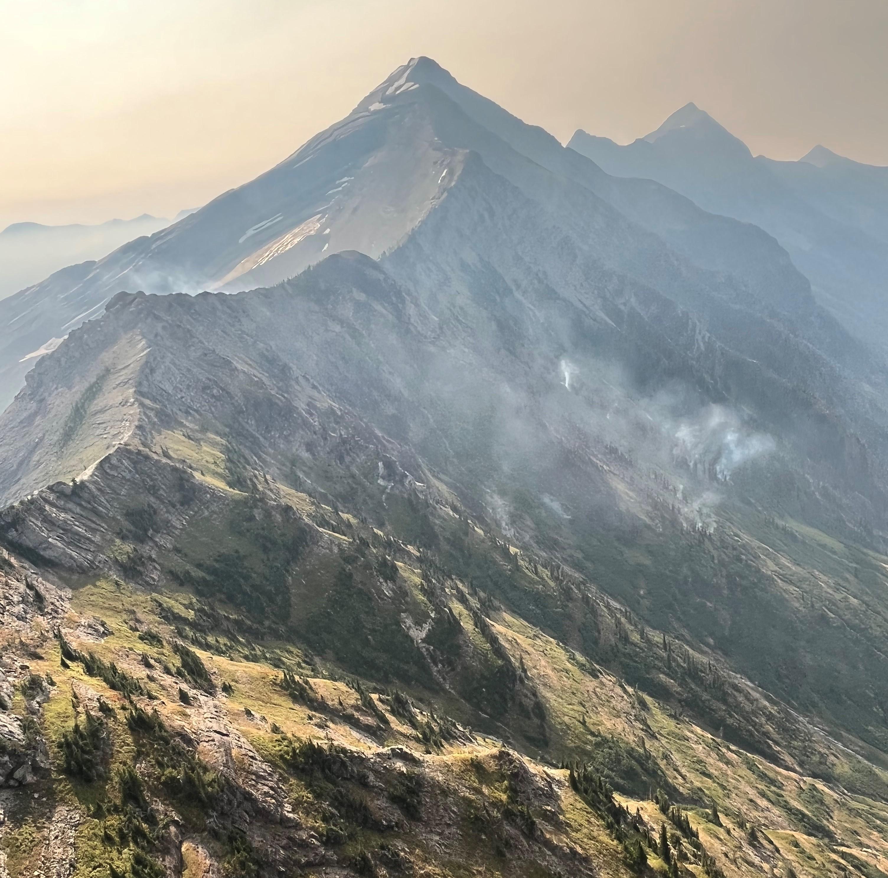 Bits of smoke rise from steep slopes along a ridge, with a rocky peak in the background and hazy skies.