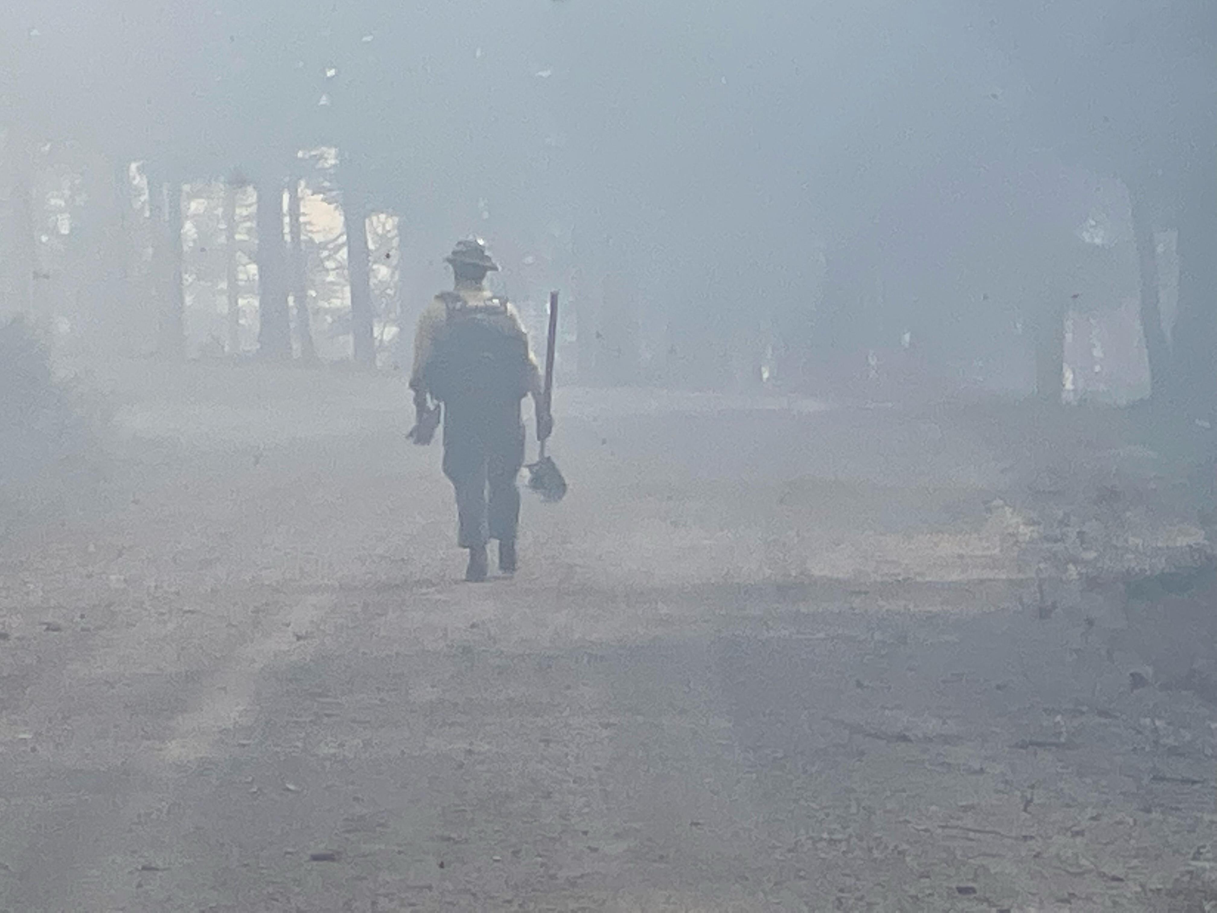 A wildland firefighter dressed in a yellow shirt, green pants, hardhat and backpack walks down a smoky hazy forested road