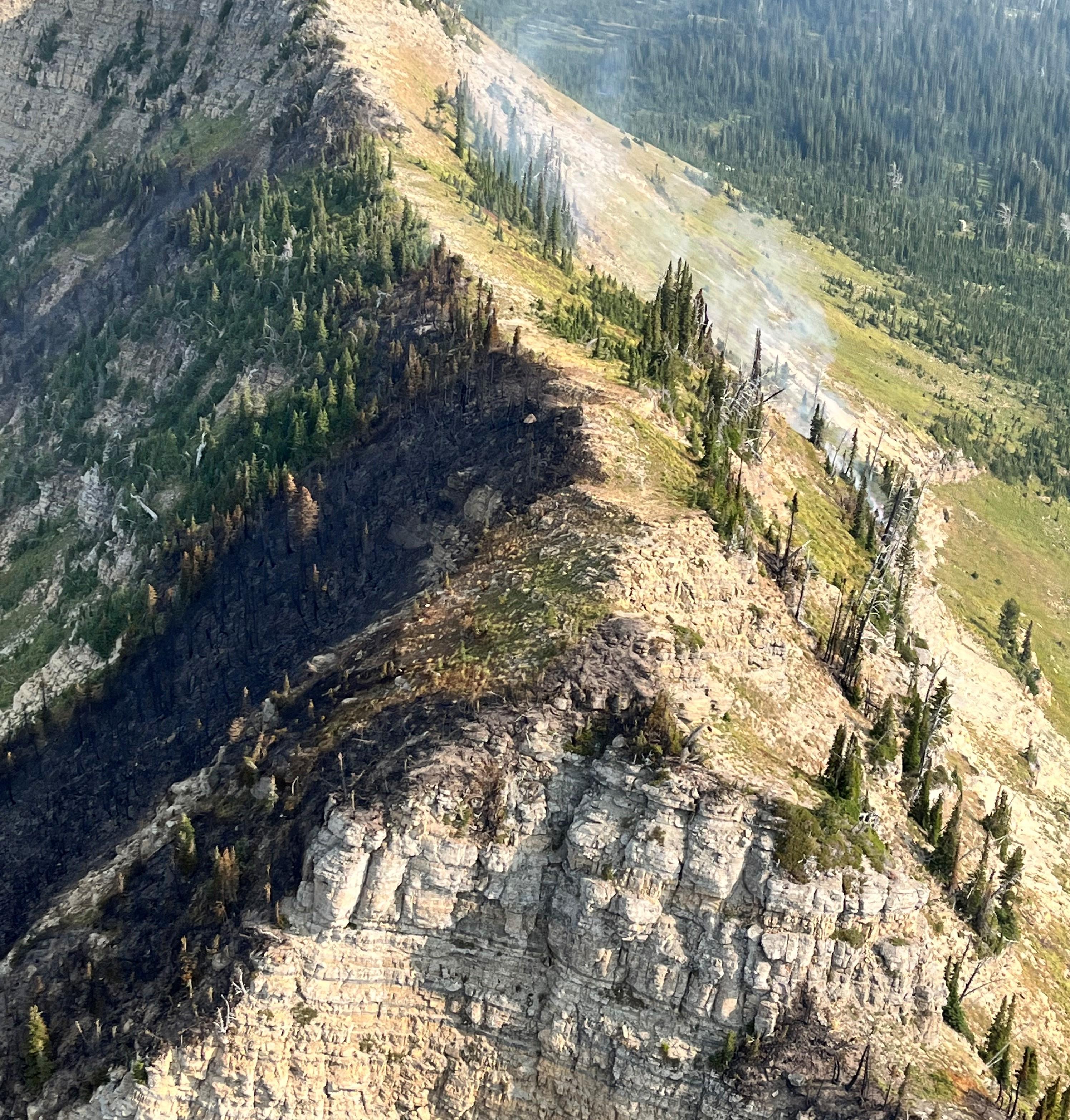 Aerial view of a rocky ridge, with a burned area on one side and a small wisp of smoke on the other side.