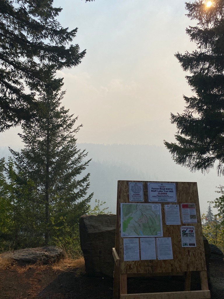 Between 2 trees is a board with information in the fore ground. Behind this there is a large quantity of hazy smoke over the scenic view of Lava falls and the Goat Rocks Wilderness Area.