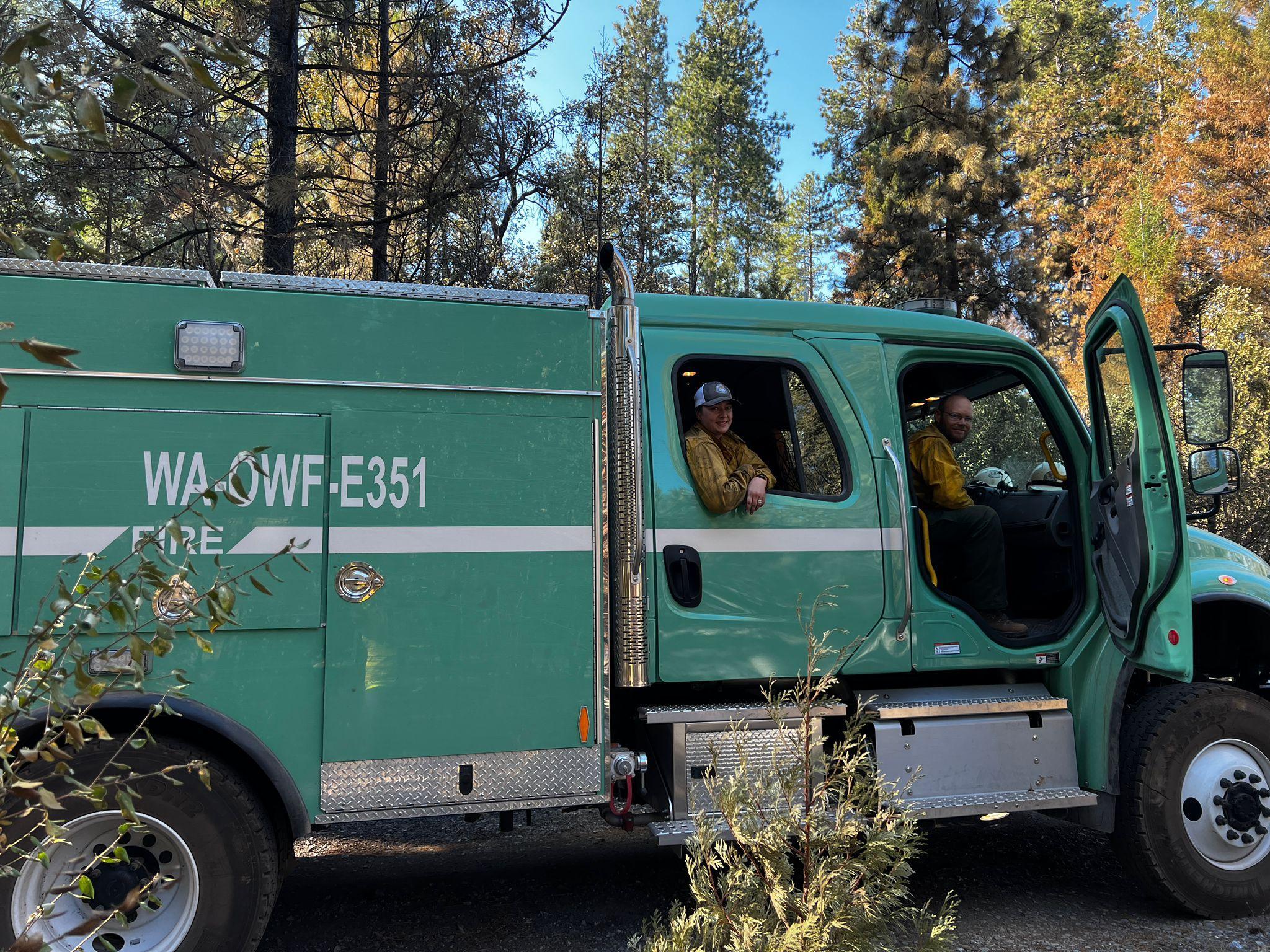 On a gravel road next to burned trees, a male and a female firefighter smile for the photographer from their green Forest Service firefighting engine