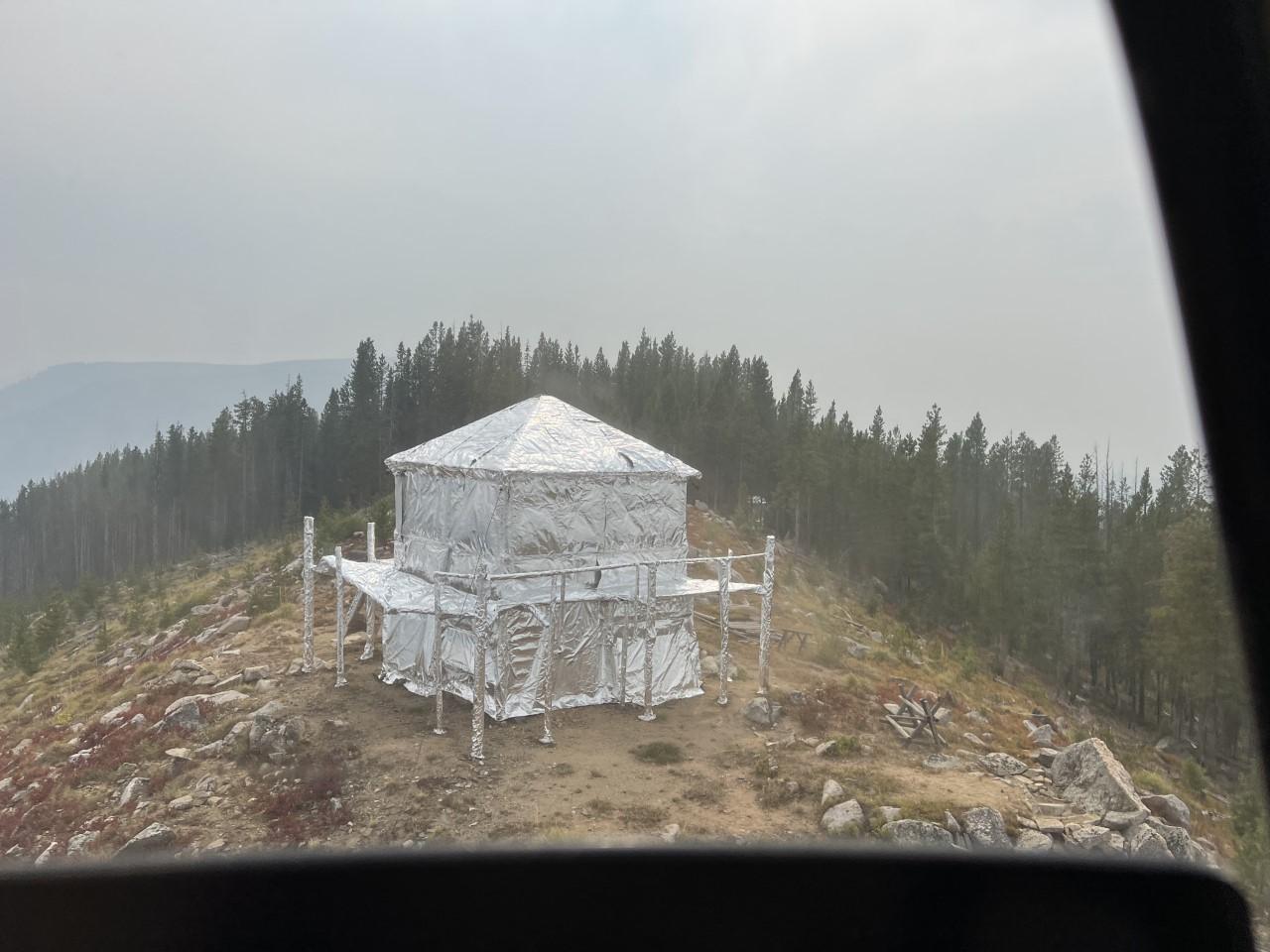 The photo is taken at the top of a mountain peak. It shows a conifer forest with a cleared area around a lookout structure that is wrapped with reflective material.