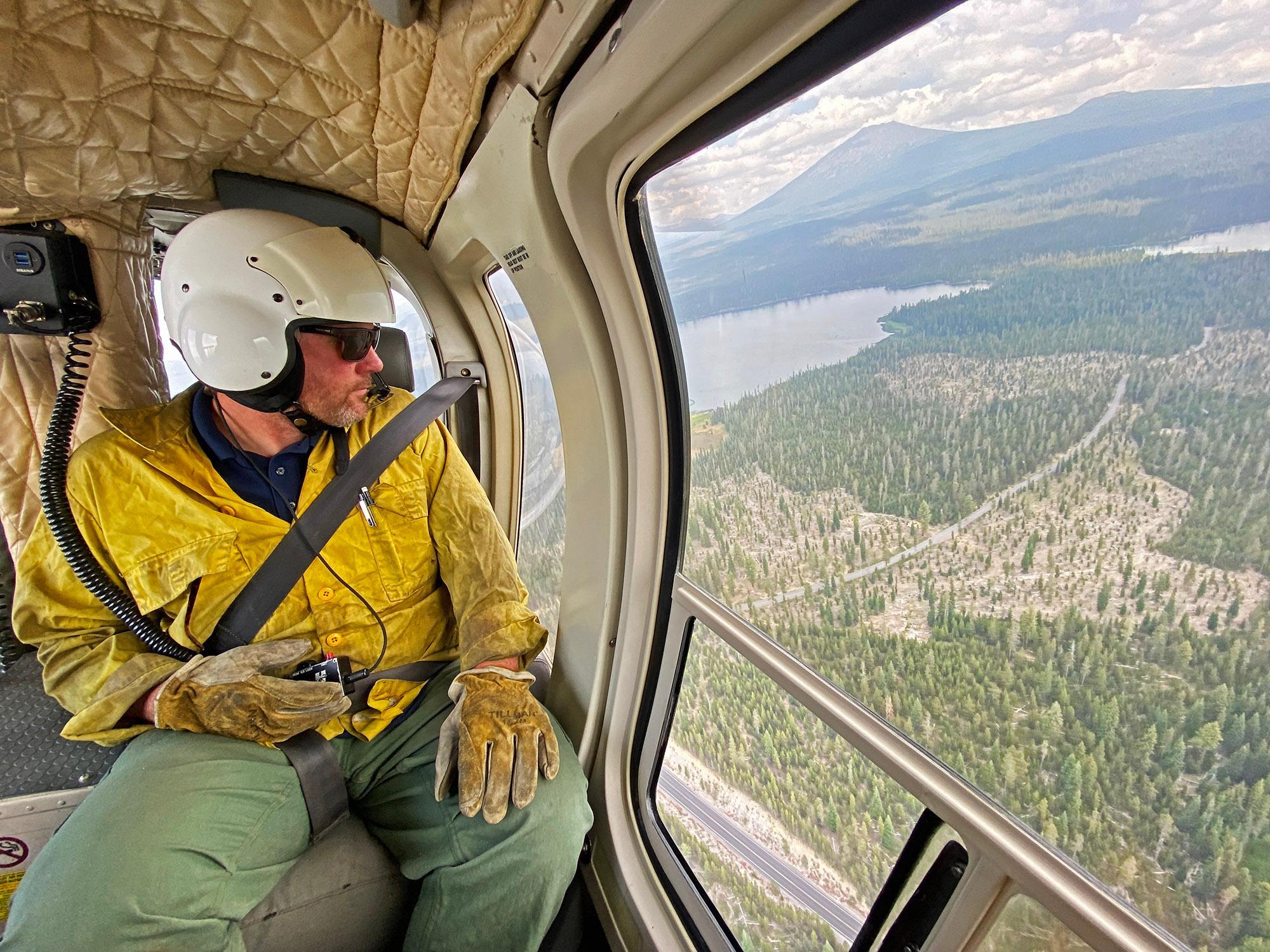 A firefighter with a white aviation helmet is sitting in the back of a helicopter looking out the window.