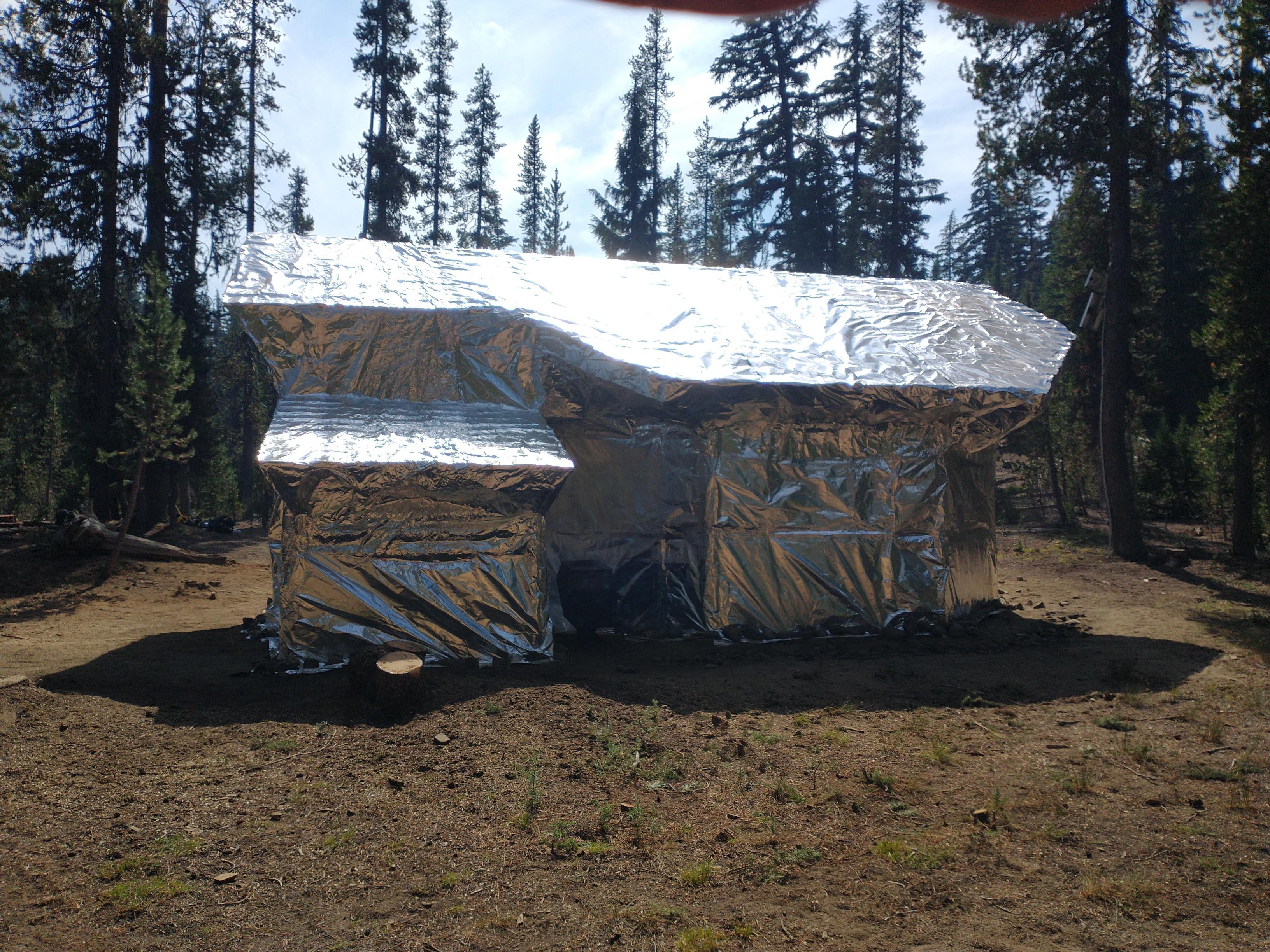 A cabin is wrapped in shiny silver foil in a pine forest