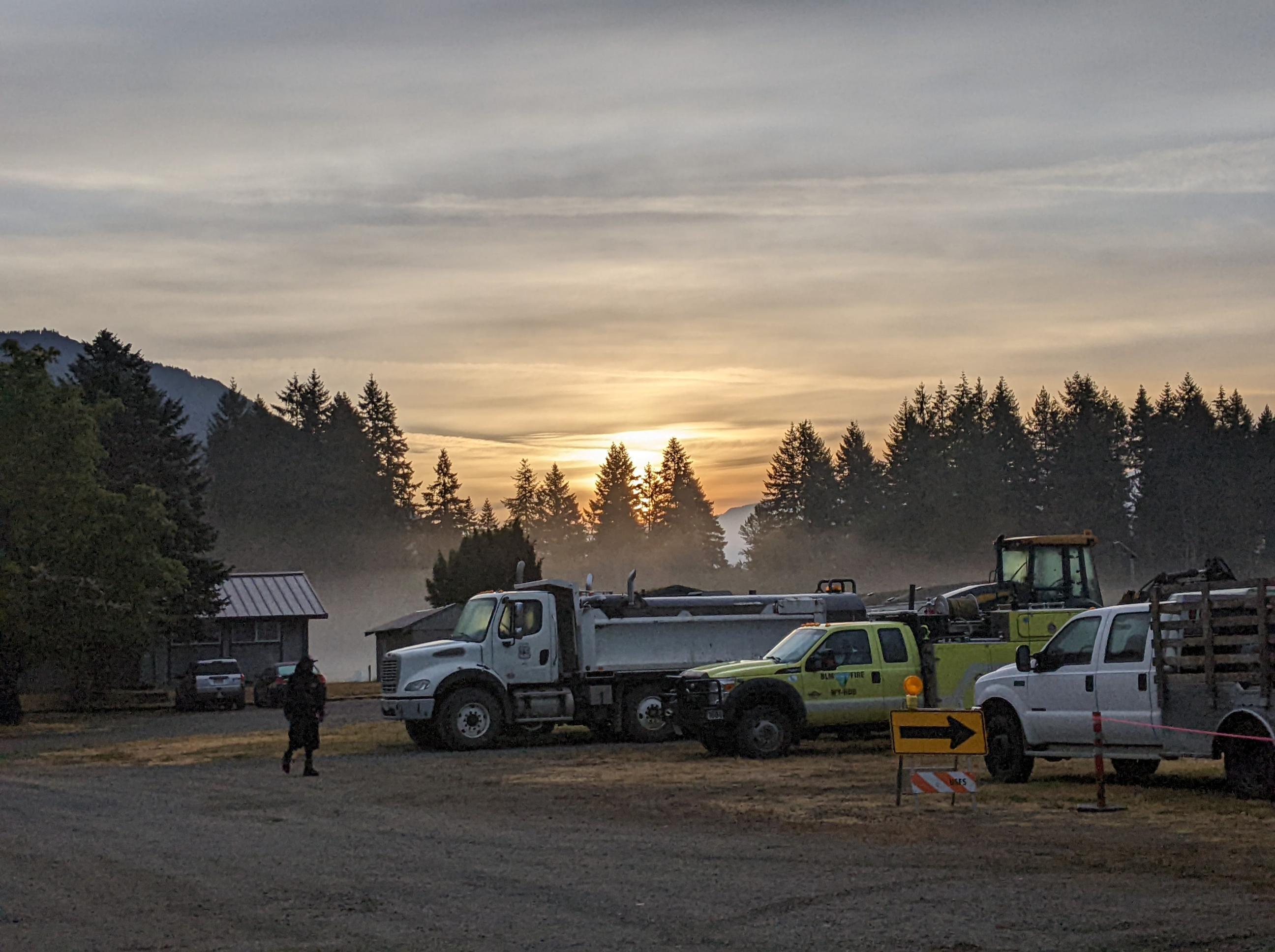The Sun rises behind a USFS dump truck and a BLM Engine. Their is a firefighter walking towards the trucks and the sun. Behind the trucks are some firs that the sun is rising over,.