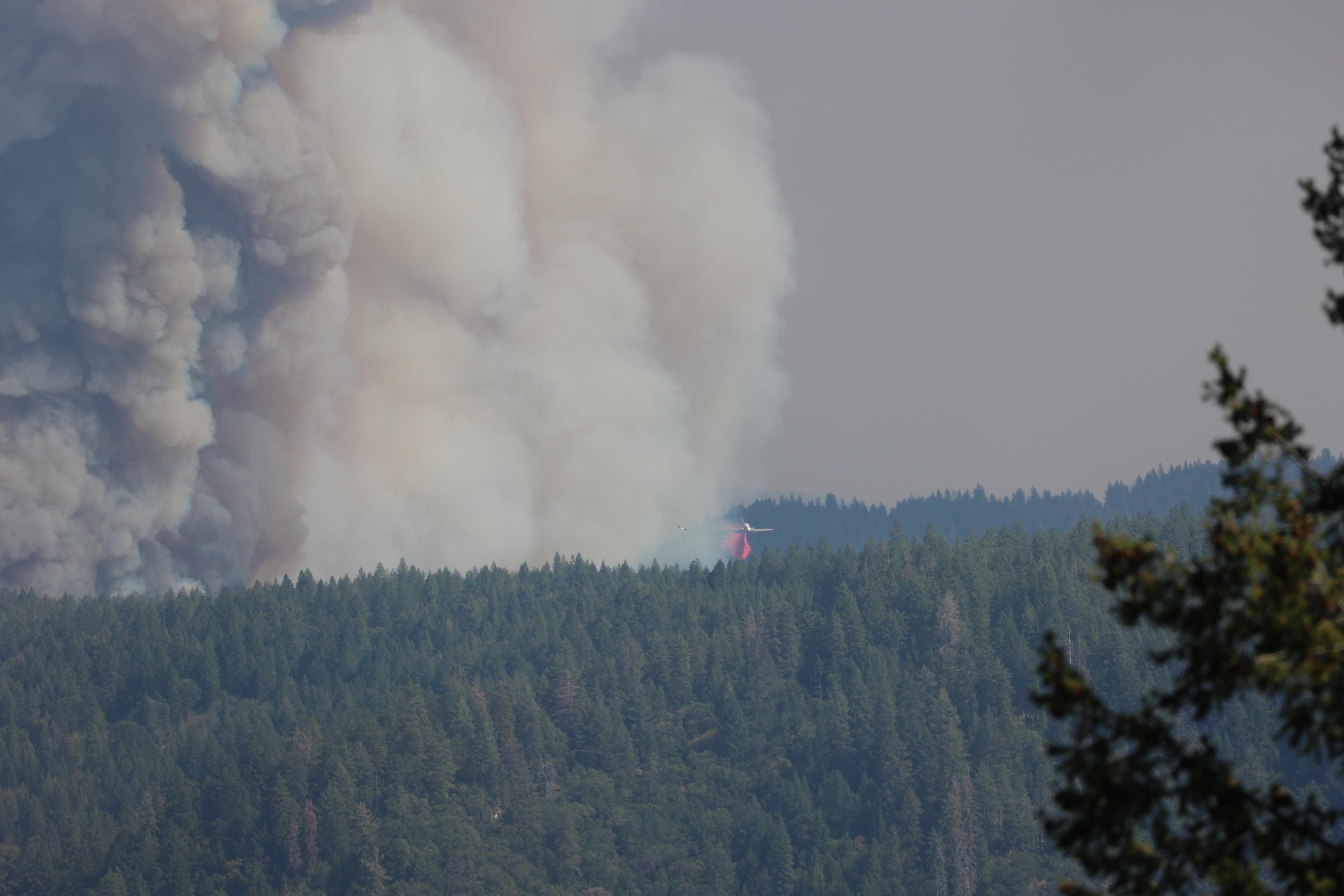 large column of dark smoke rising far into the sky as the fire burns coniferous mountain slope. an air tanker drops pink retardant on the edge of thick smoke