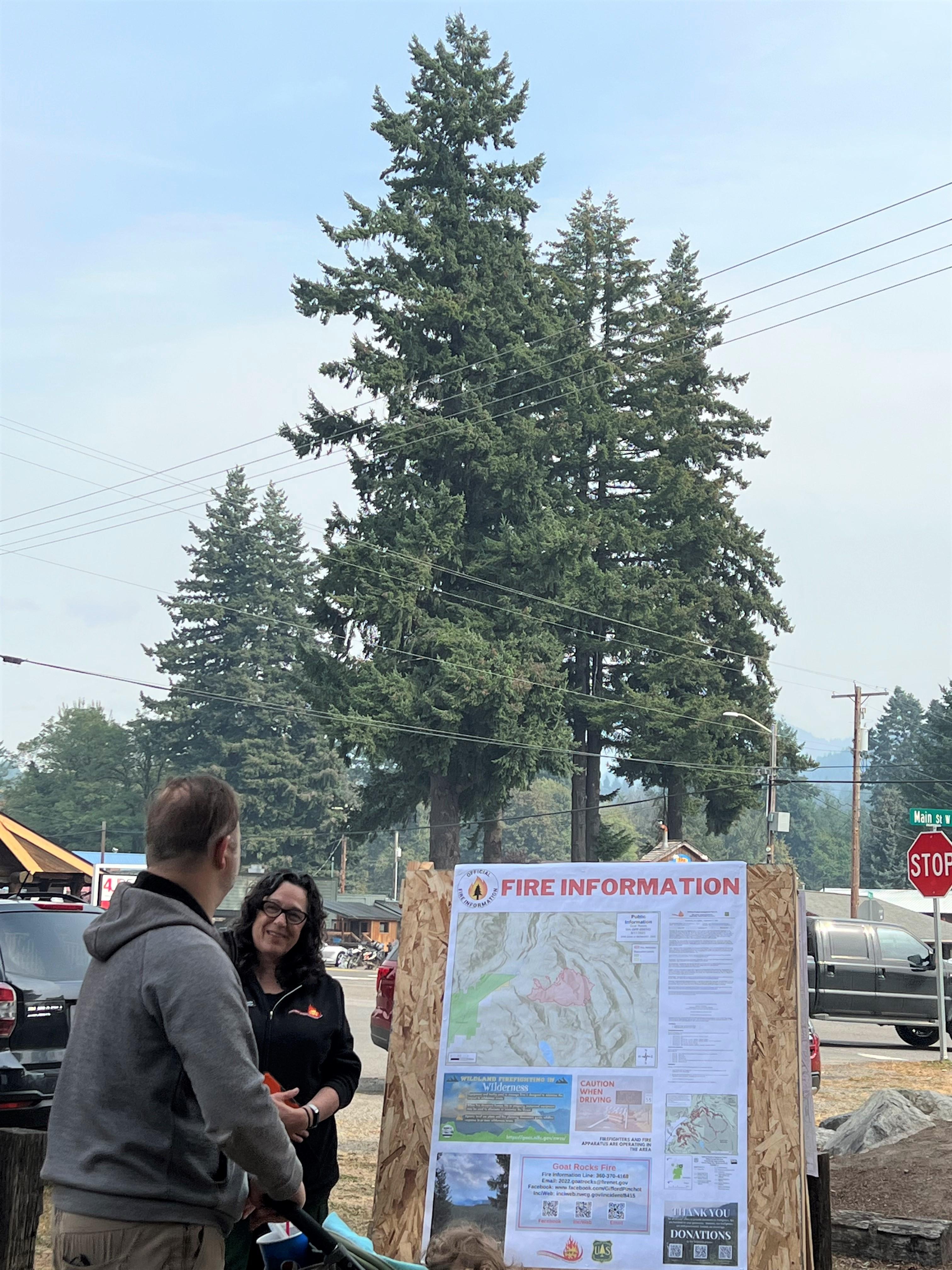 In the background stands a large Fir tree. In front of that is a plywood sandwich board with a poster full of information and a map on it. Next to it are 2 people, one is a public information officer and the other is a member of the public asking the PIO.