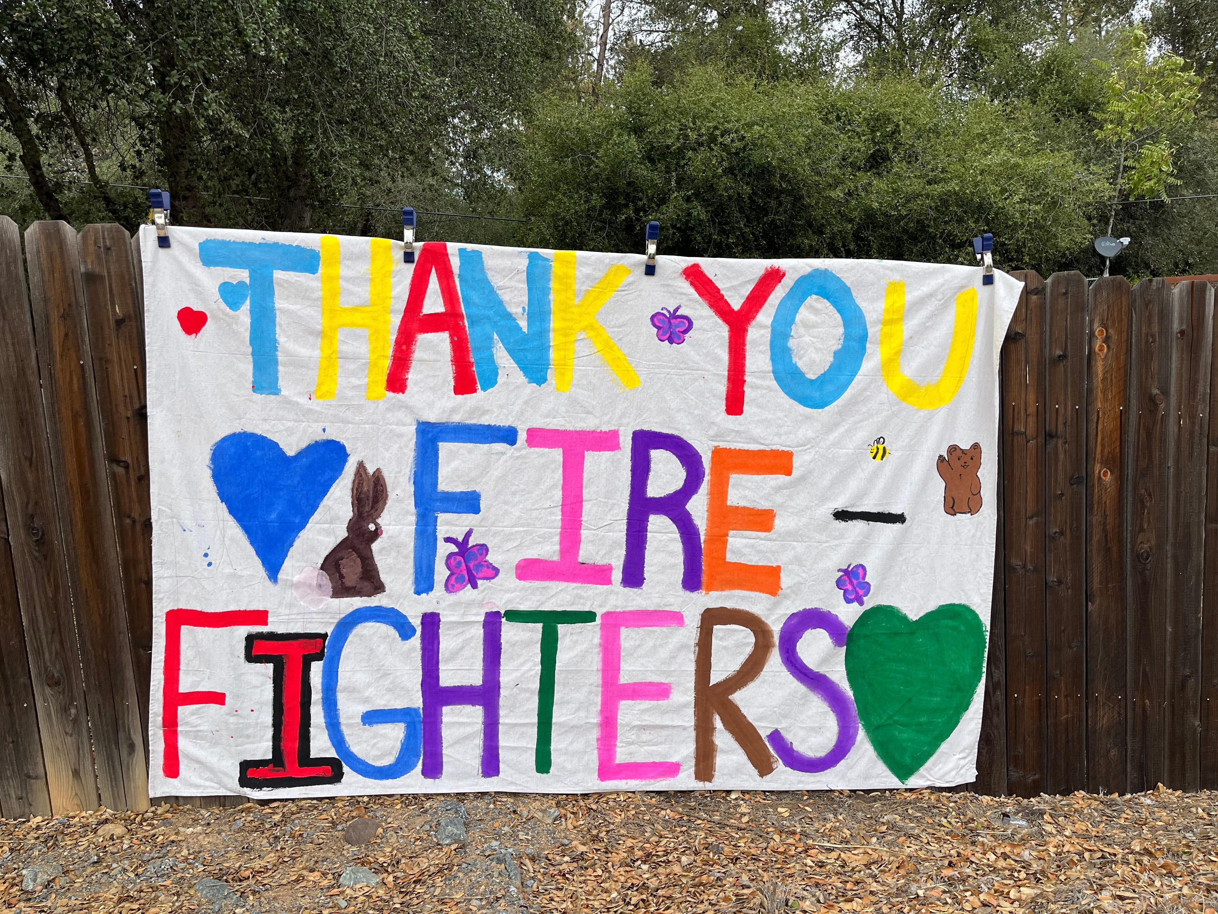 Thank You Firefighters says a colorful hand-painted sign hanging on a fence for firefighters to see