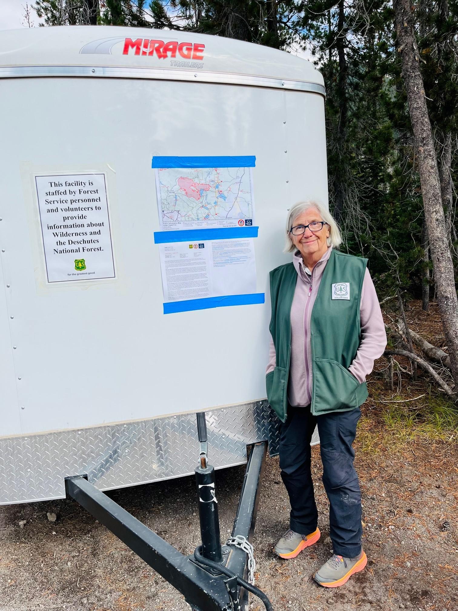 A woman stands to the right of a map, which is taped to the front of a pull behind trailer.  She is wearing a vest with the US Forest Service logo on it, indicating that she is a volunteer.