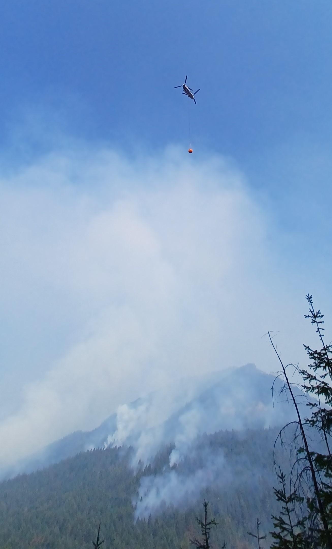  A helicopter with a large bucket of water hanging below it flies over the fire area. Smoke rises through the trees on the forested slopes.