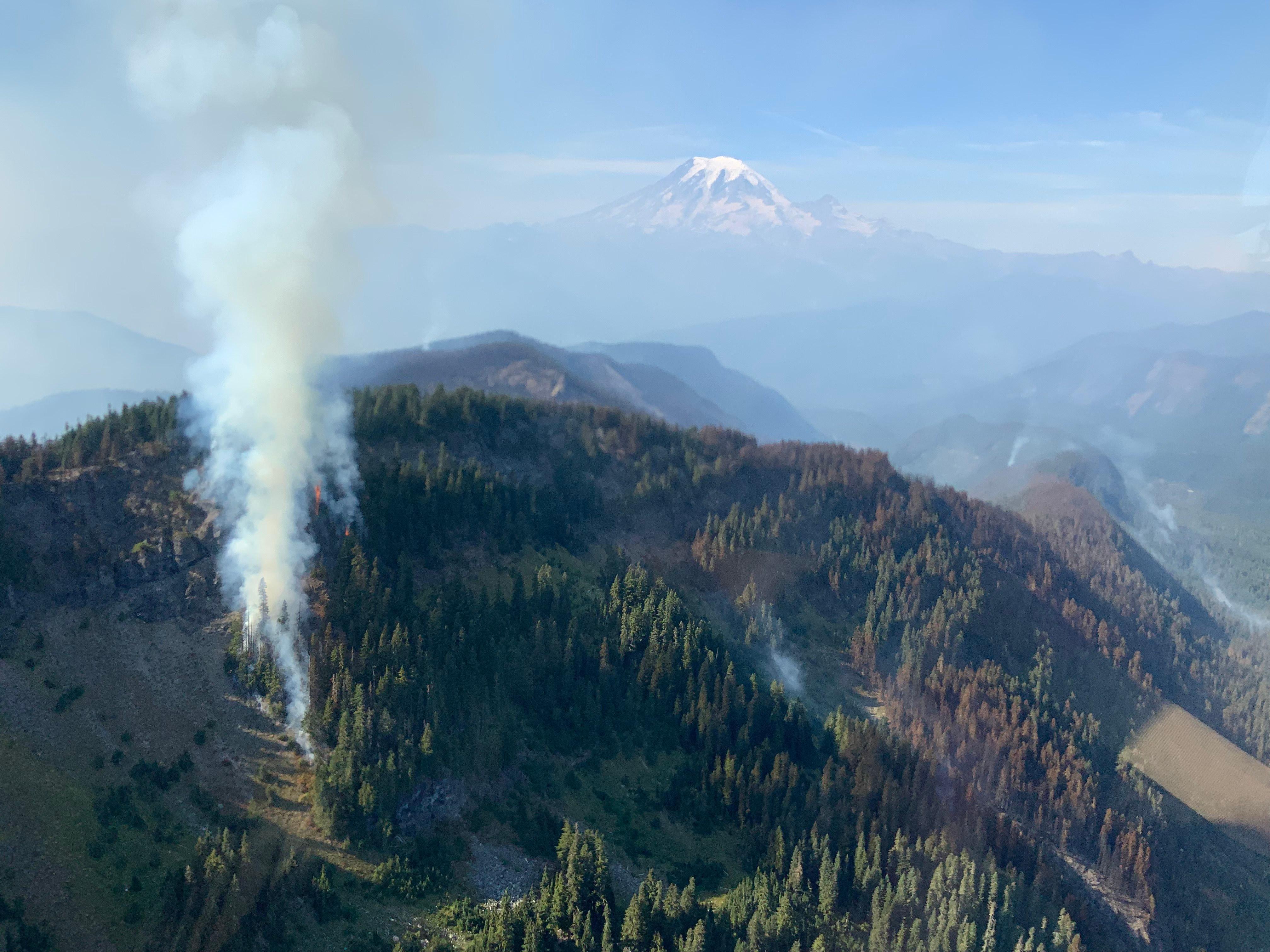 A photo taken from a helicopter looking down on part of the Goat Rocks Fire. Smoke is rising from a few patches of the forested areas of a steep knob, with many rocky areas showing. Mount Rainier is visible in the distance.