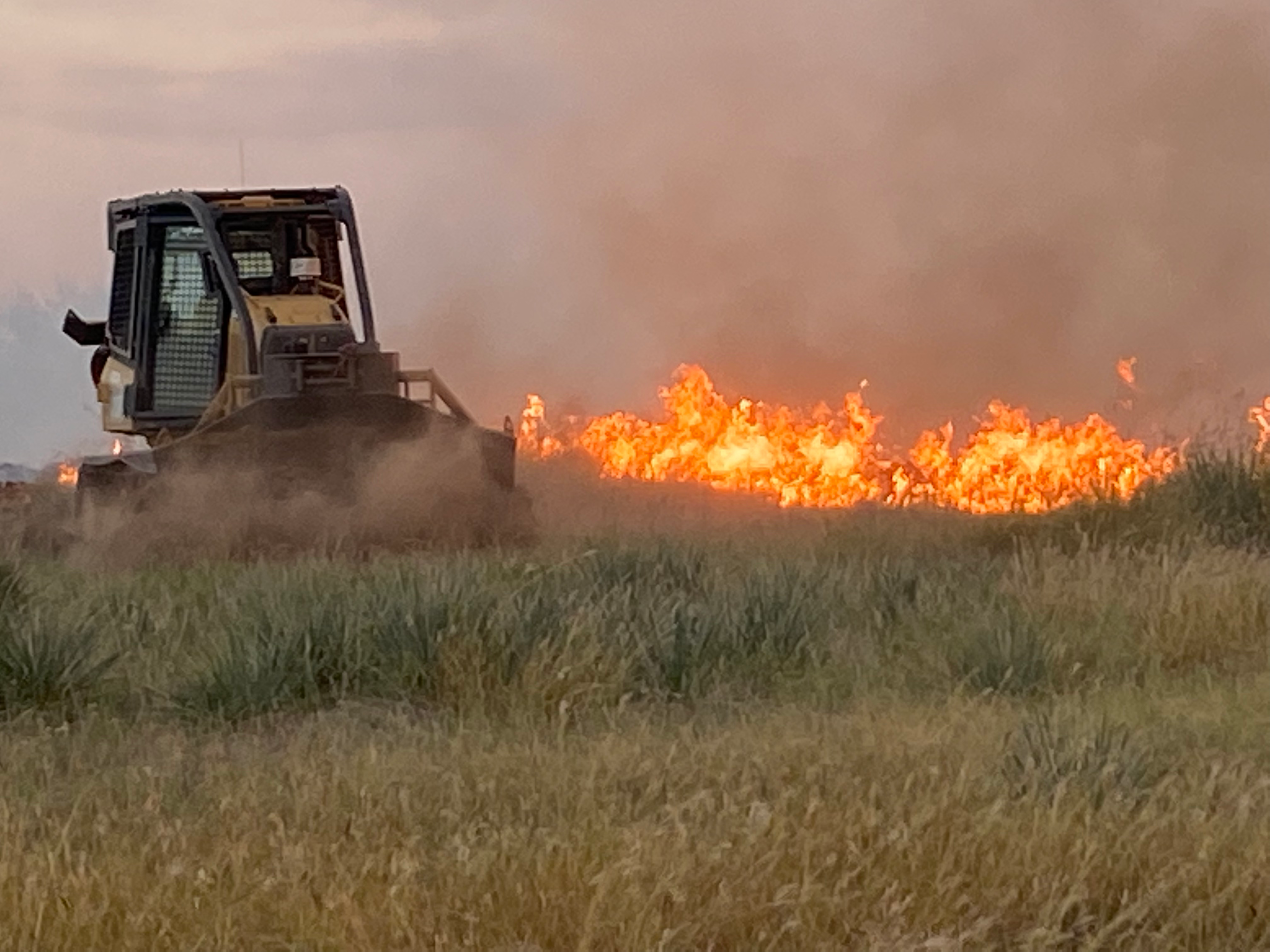 Dozer is seen pushing dirt and creating a fire break from left to right  and visible flames  on the other side of the dozer