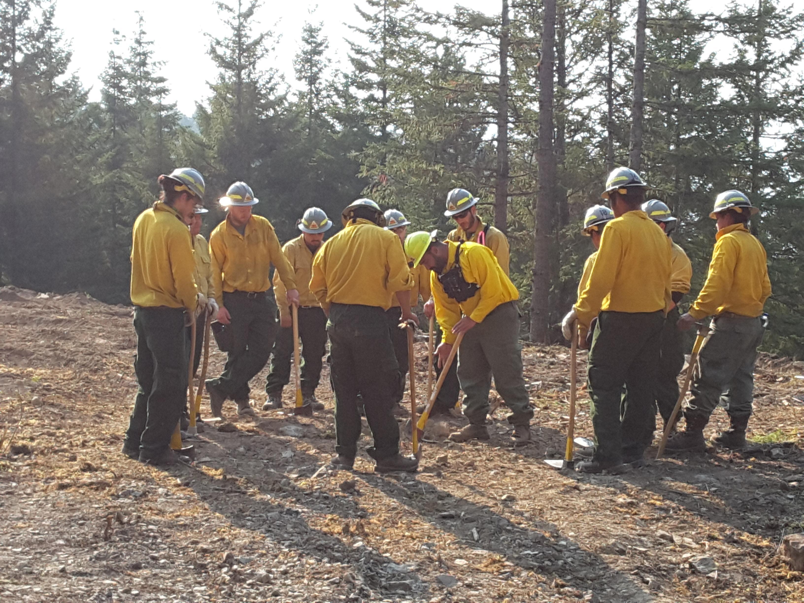 A crew of firefighters are gathered in a circle in a cleared area. Their Crew Boss is demonstrating how to use tools correctly to train newer crew members.
