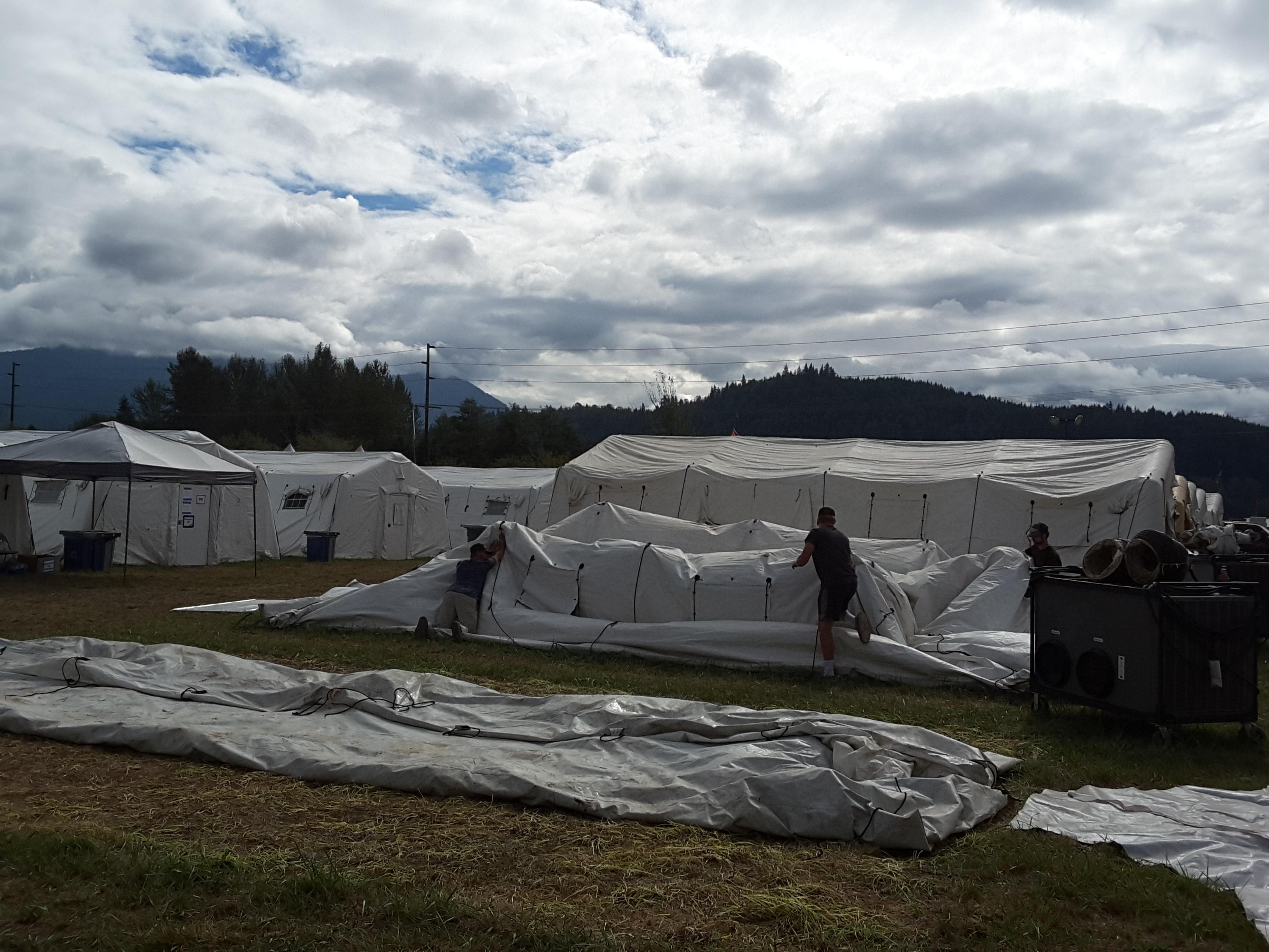 Several men are working to collapse a large yurt so it can be folded and hauled away. A flattened yurt is in the foreground. Several standing yurts are in the background.