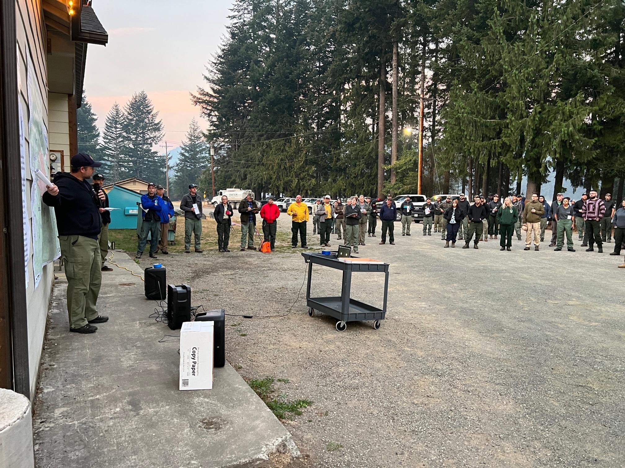 A group of people are standing in a large semi-circle, watching a man who is discussing the fire and pointing at a map on the side of the Packwood Community Hall.