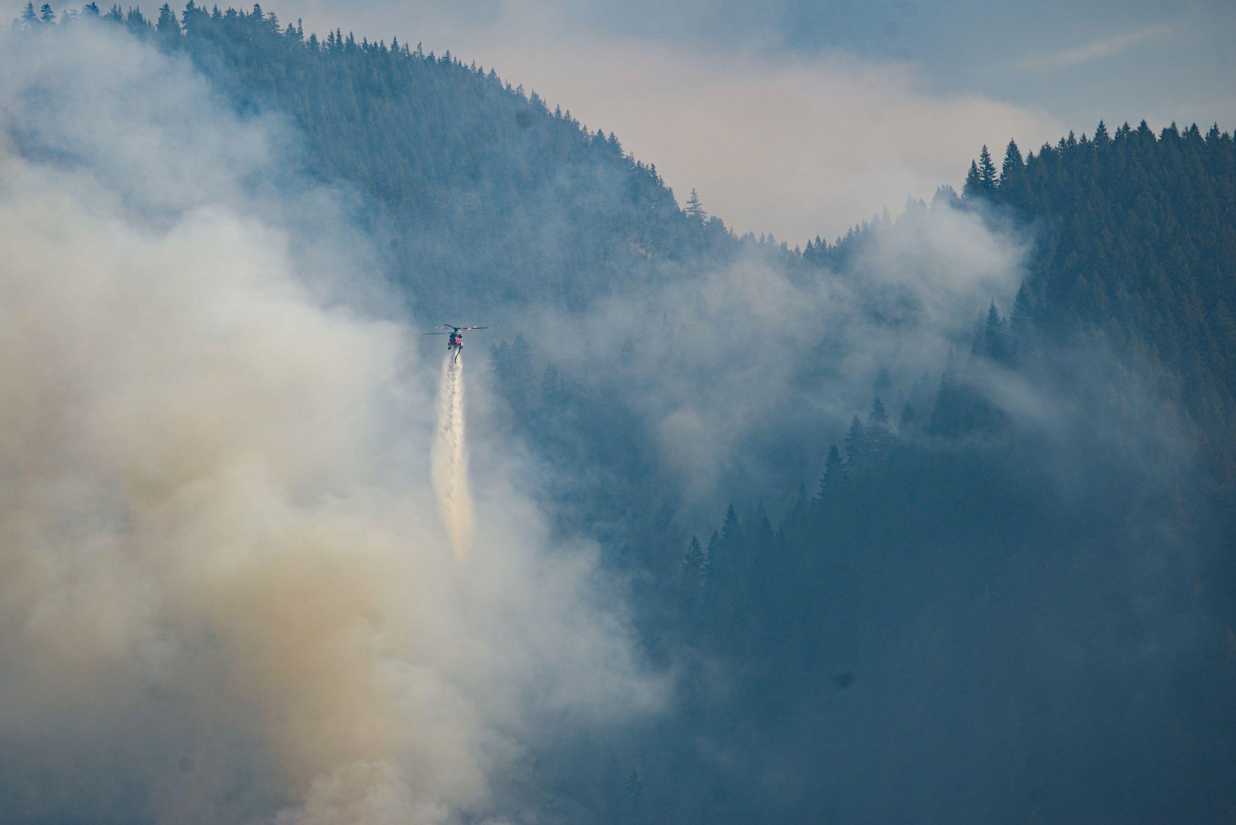 A helicopter is dropping water on the burning forest south of Coal Creek. Smoke rises from the forest on steep slopes below and behind the helicopter.