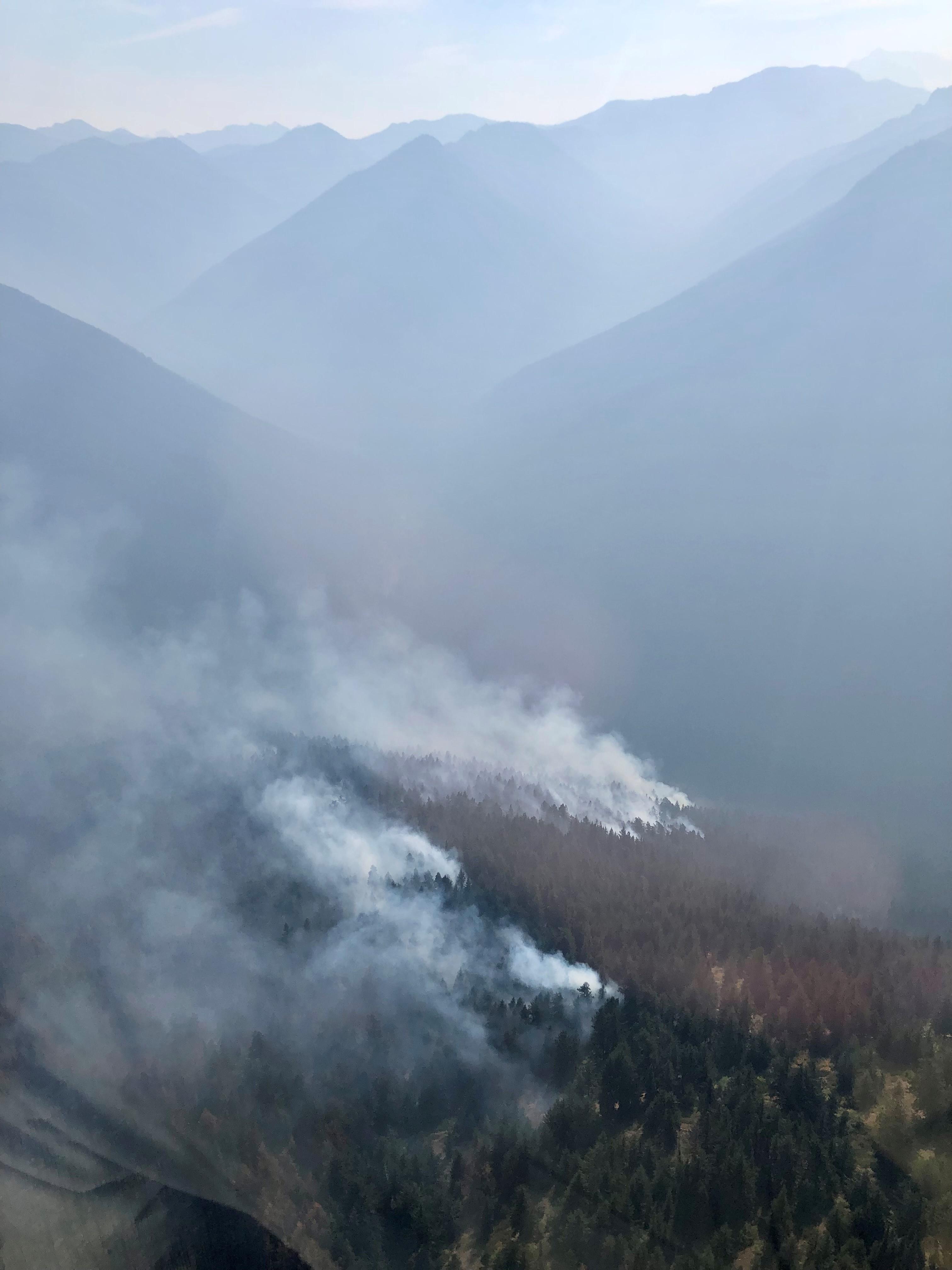 An aerial view of the Three Fools Fire on October 4, 2022. Smoke is drifting near the tree tops and filling the valley behind in haze.