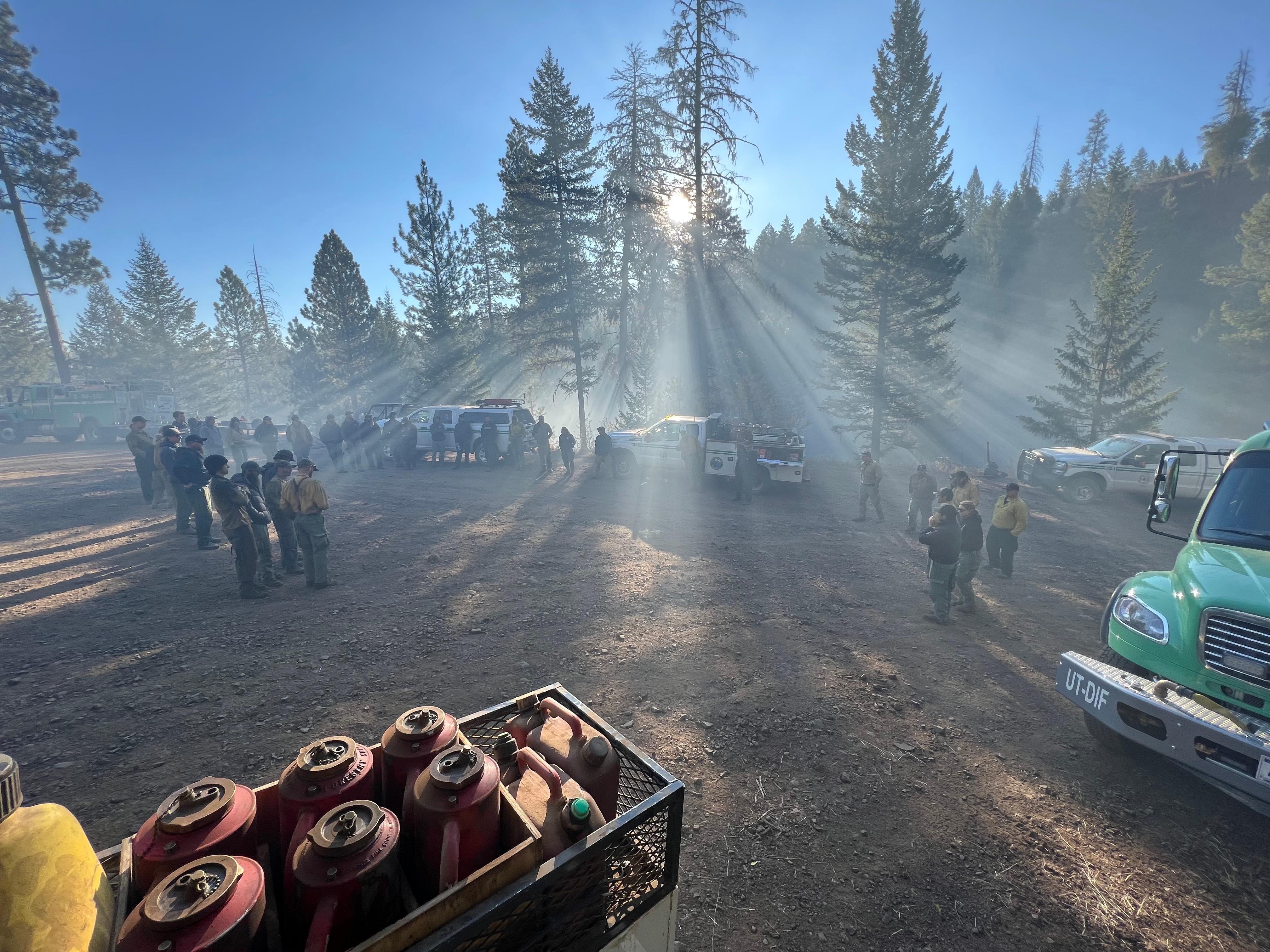 Firefighters are shown gathered around vehicles as the sun comes up behind them. Light smoke in the air highlights the suns’ rays through breaks in the trees behind the firefighters.