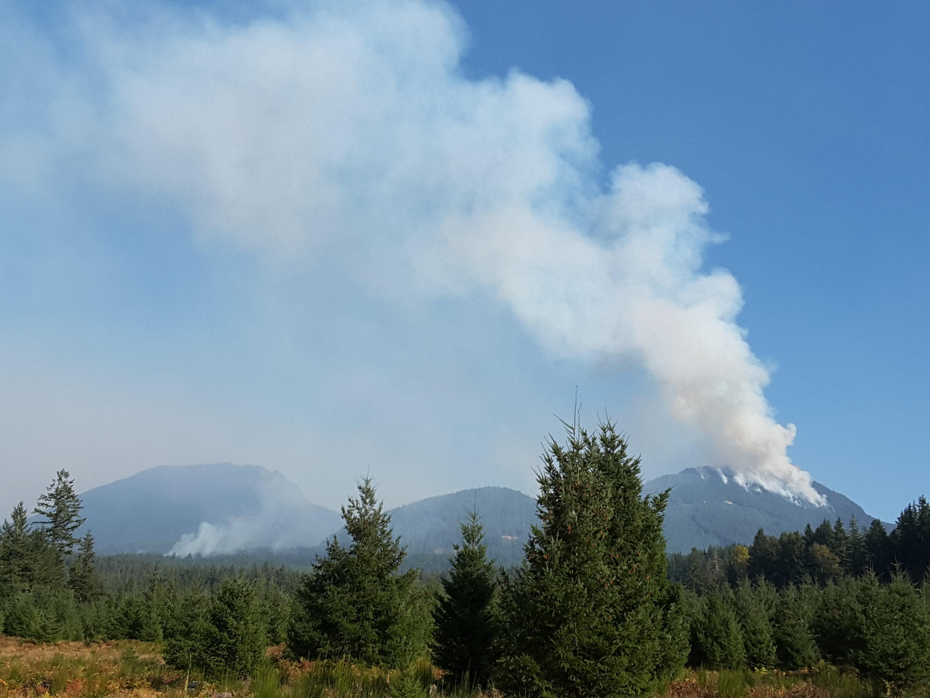 Smoke from the Goat Rocks Fire rises from a steep, forested slope near Three Peaks ridge on the right side of the photo. A lesser amount of smoke rises from the forest near the left side of the photo, in the Coal Creek drainage.
