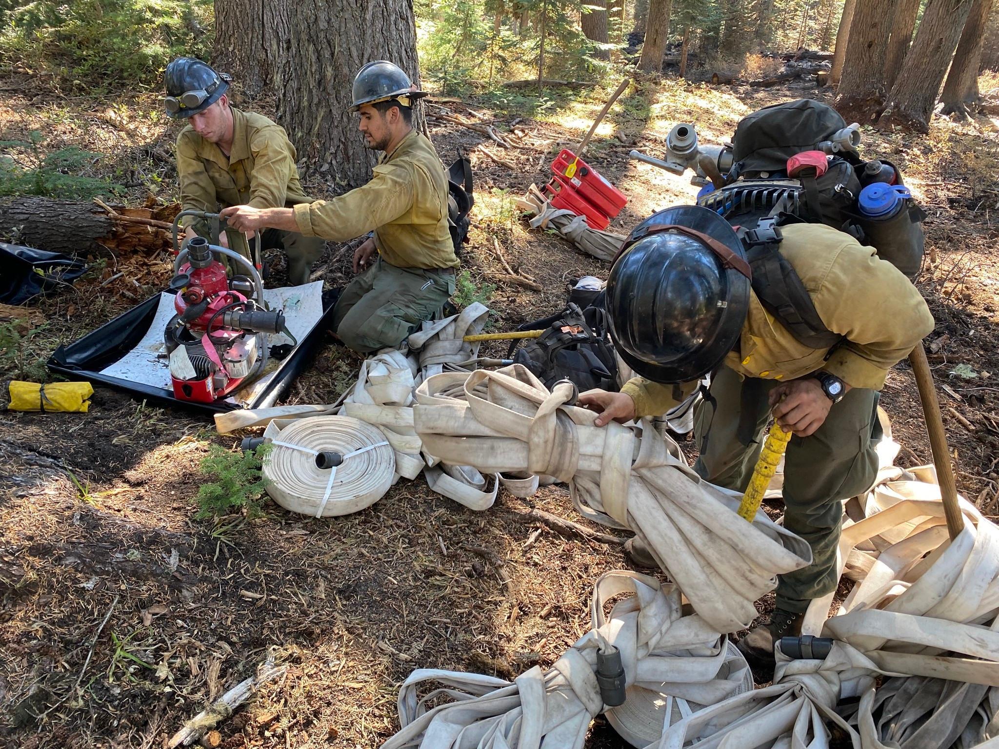Three firefighters in black hardhats, yellow shirts, and green pants cleaning up equipment in the equipment. Equipment includes a red a silver metal pump use for water and numerous loops of white fire hose.