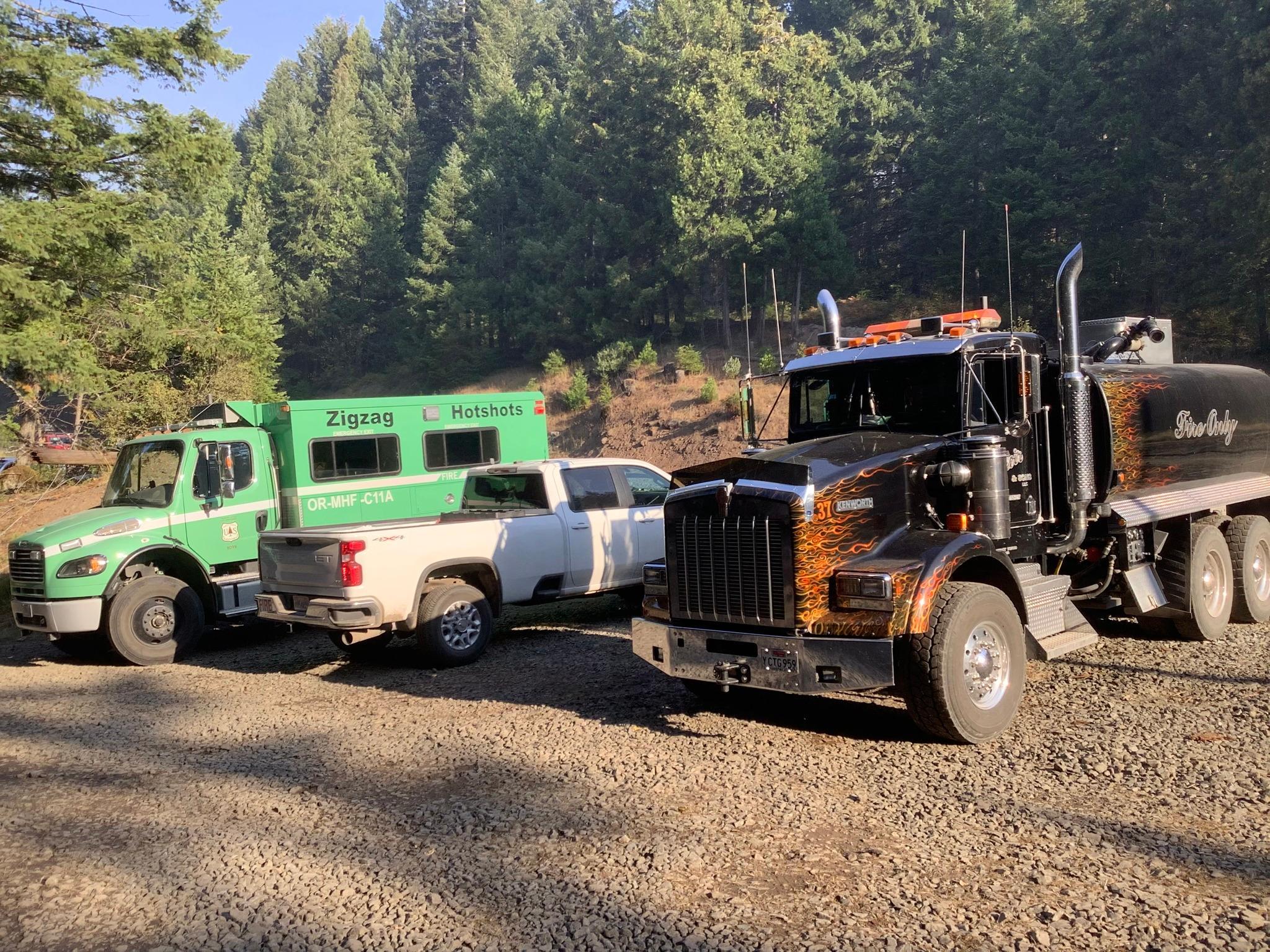 Vehicles parked in a gravel parking lot surround by trees.  Vehicles are a green buggy transport, white pickup truck, and a black water tender.
