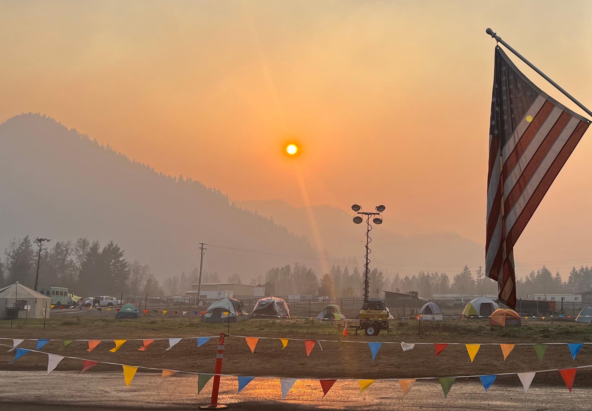 View from the command post has American flag in the foreground, camping tents and vehicles while the orange sun rises above the mountains and smokey haze in the air.