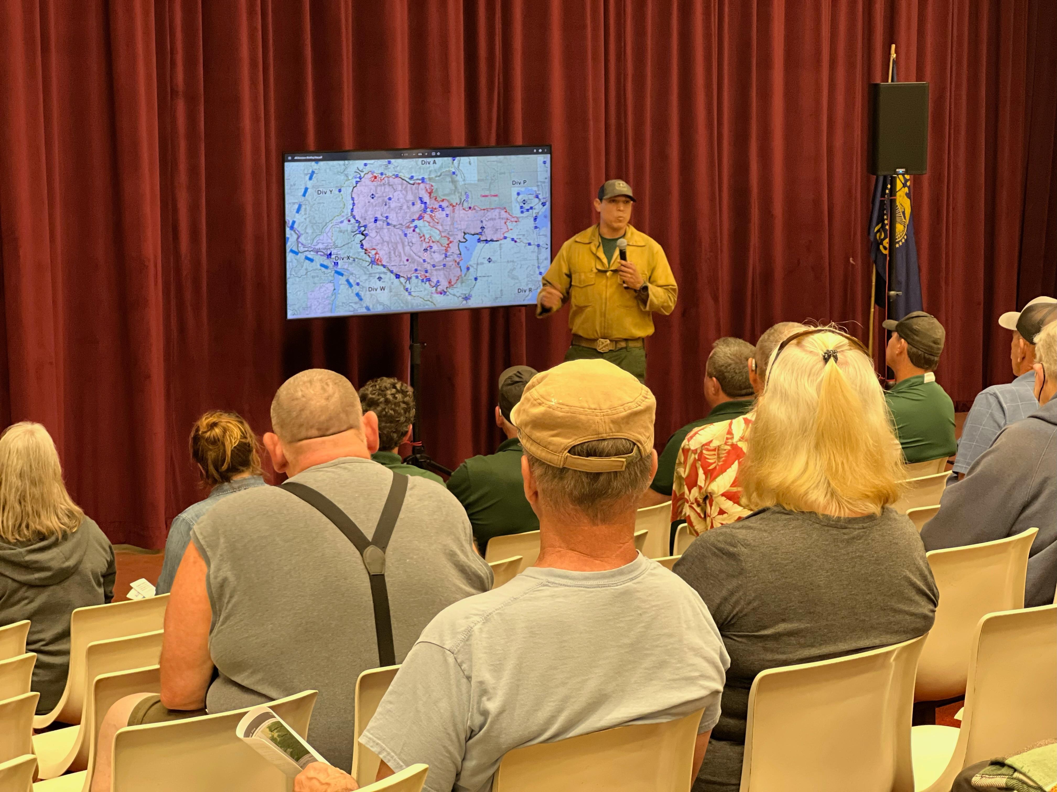 People sit in chairs looking at a man in a yellow shirt who is presenting fire information next to a tv screen.