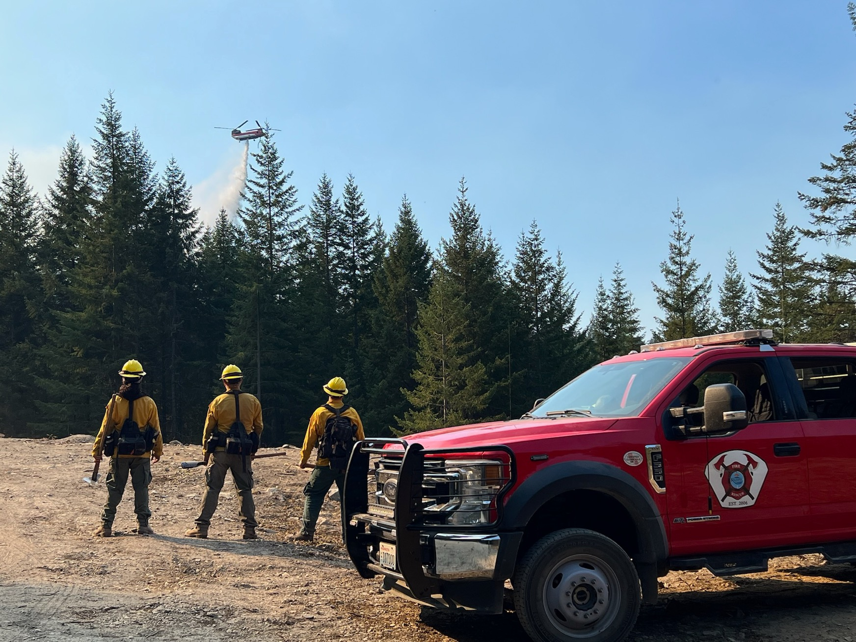 Three firefighters stand by their truck along a road. They are  watching a large Chinook helicopter drop water in the Coal Creek drainage in the background .
