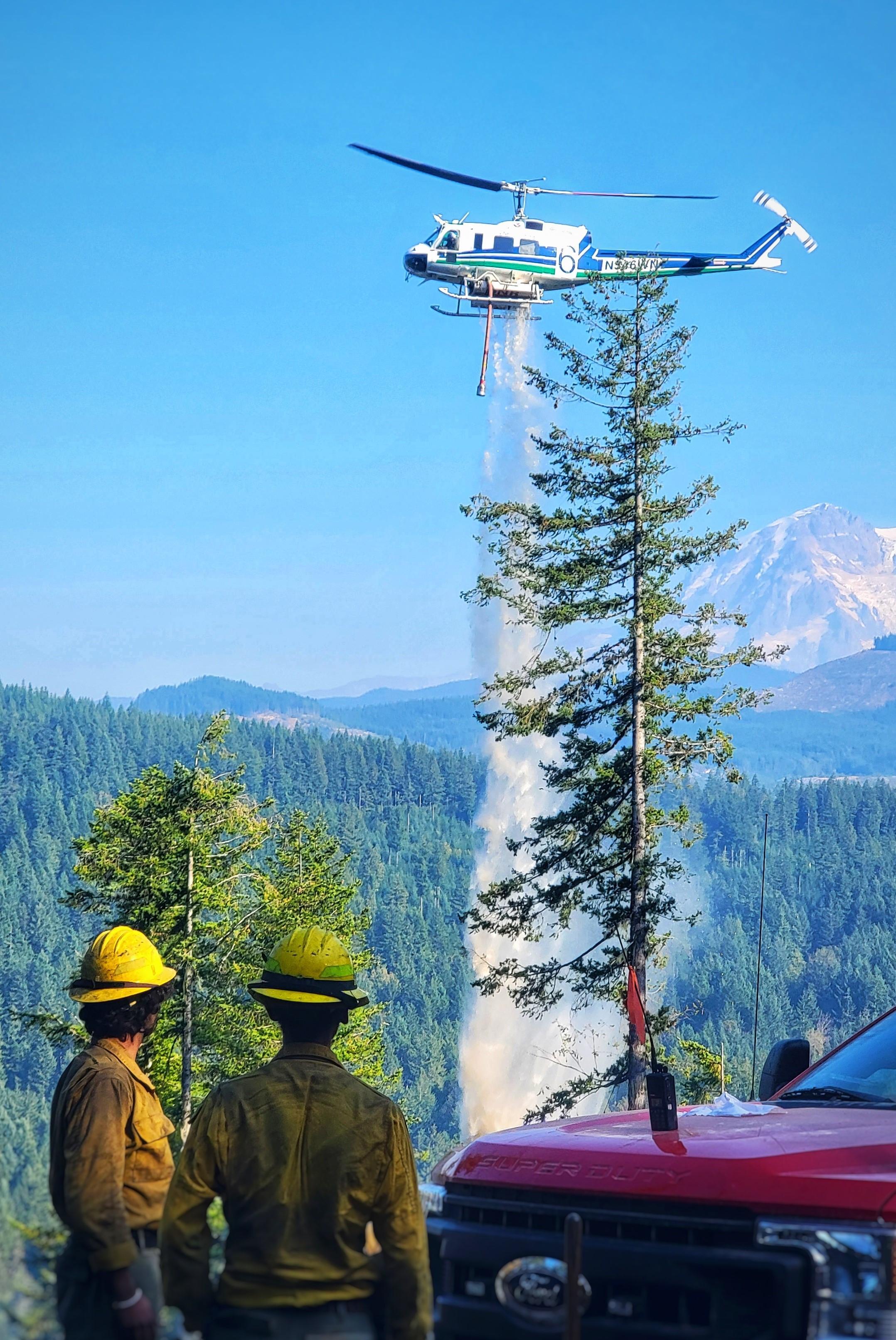 The south side of Mount Rainier looms in the background as firefighters in wildland gear stand beside a red truck, watching from a safe distance as a helicopter drops water onto the eastern perimeter of the 8 Road Fire in the afternoon on 10/18/22..