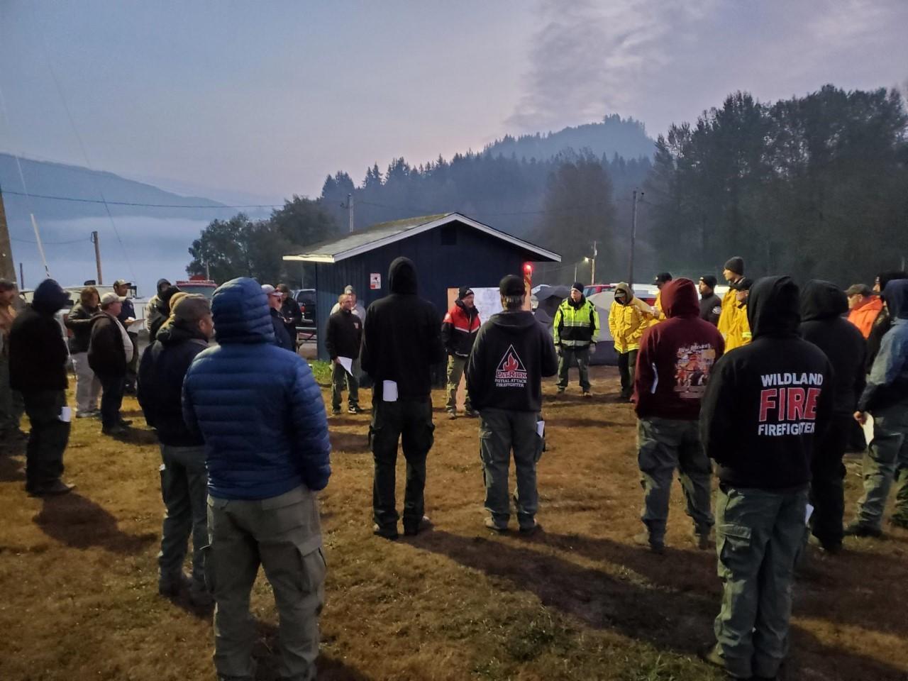 Incident commander briefs fire crews at the beginning of their shifts. Sun is just rising and mist/smoke is visible over the background which includes a small lake, tents and forested hills.