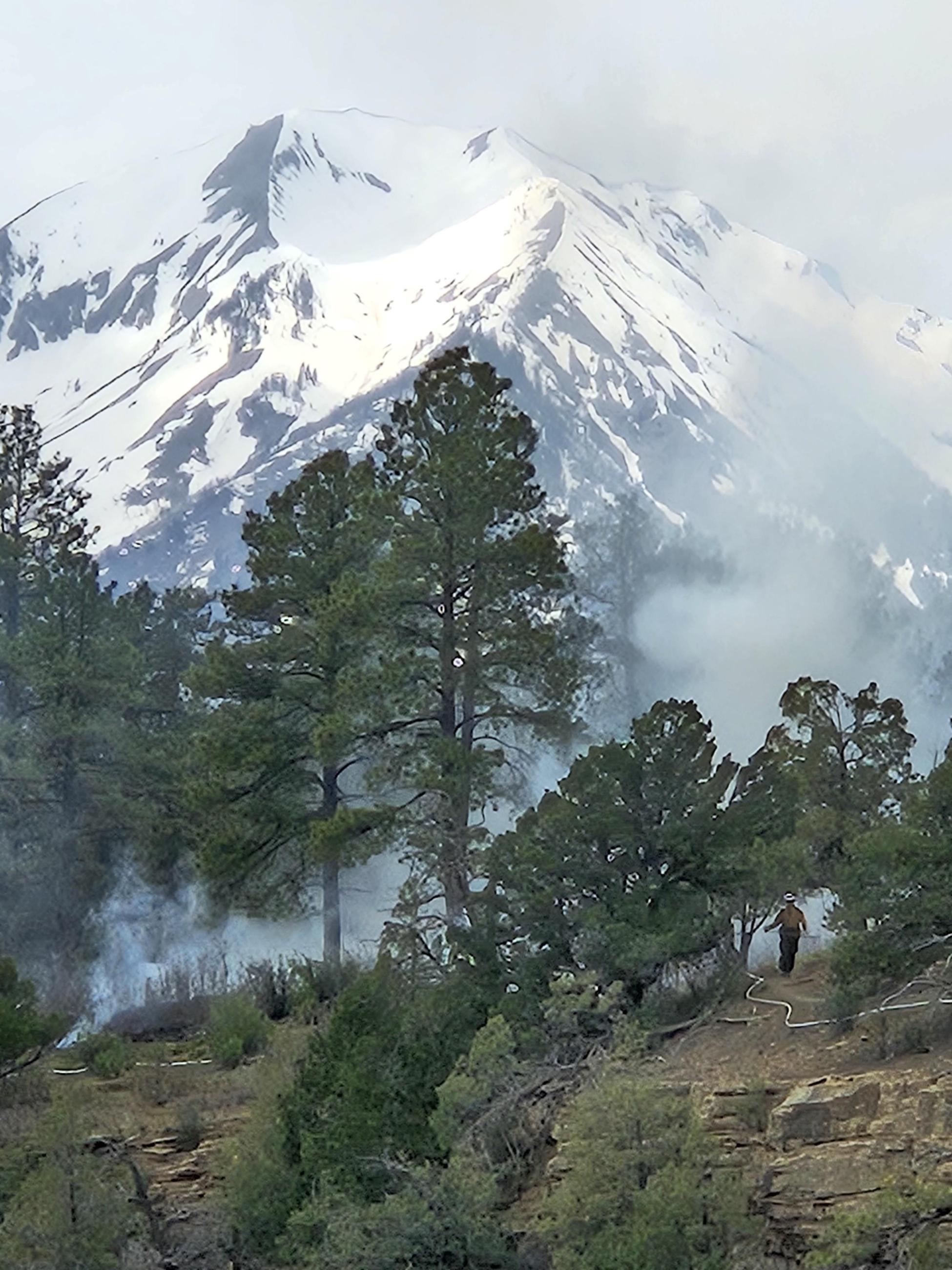 Image of firefighter walking along trail on Animas City Mountain Prescribed Burn. Image has Ponderosa pine trees, Juniper and Pinyon trees, smoke, rock, brush, fire hose and very large snow capped mountain in the background. 