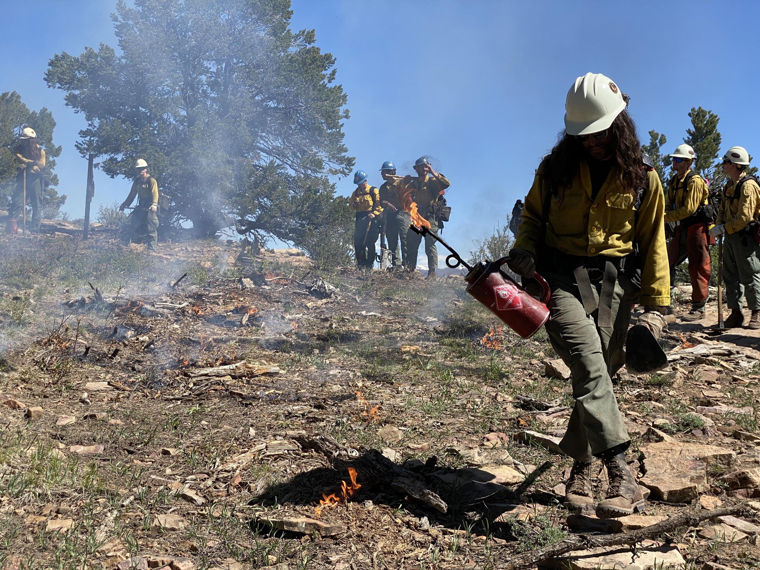 Firefighter holding a red drip torch in the foreground of the photo with a small flame coming out of tip. There are short flames and smoke in front of him. Seven firefighters holding in the background with packs on and tools in their hands, all wearing green pants, yellow shirts, and hardhats. 