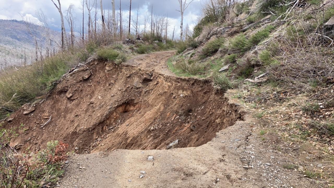 Image shows a gravel forest road with a portion of road missing due to erosion and storm damage.