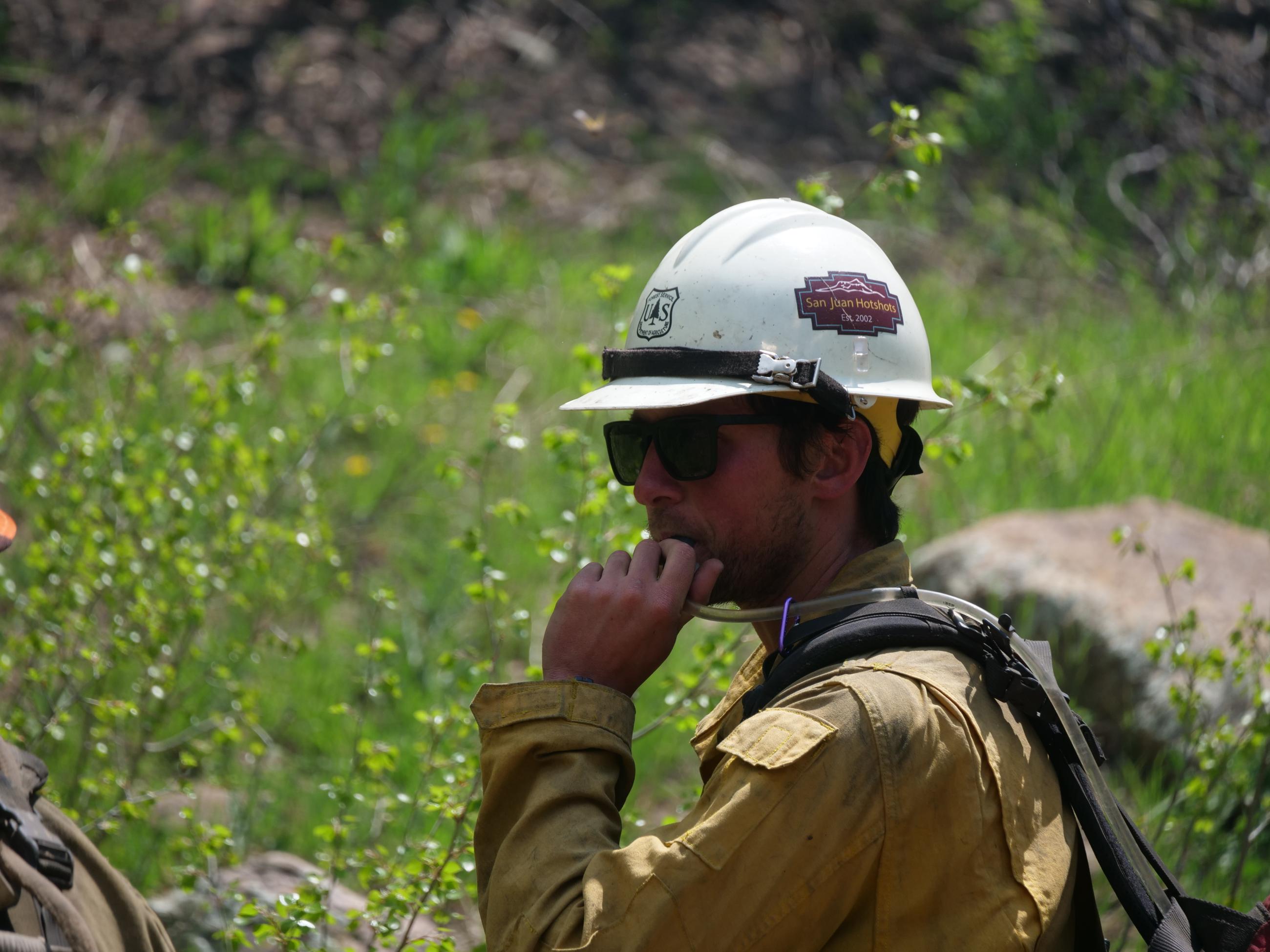 A firefighter in a white helmet with the words "San Juan Hot Shots" is seen taking a drink from his hydration pack.