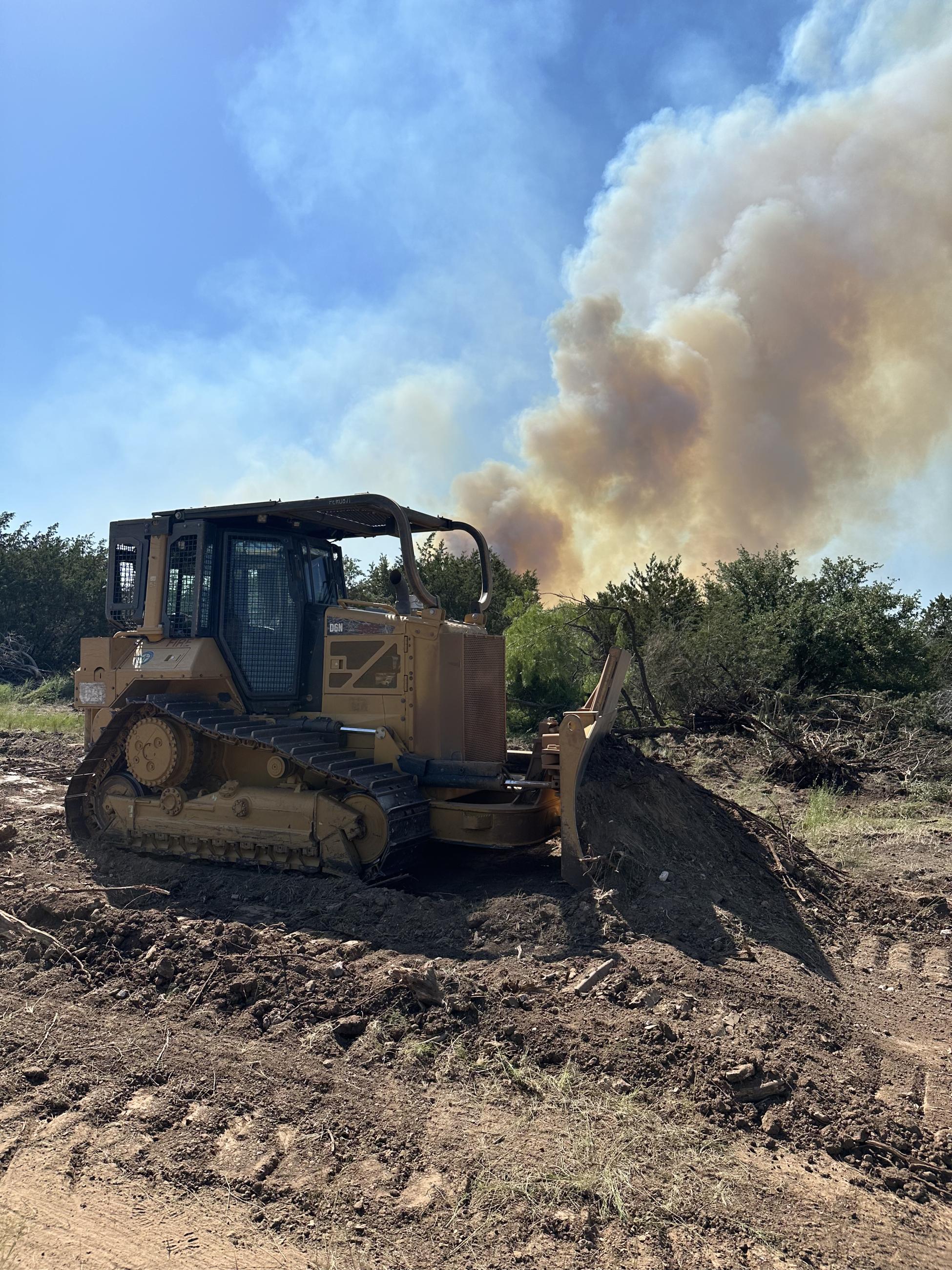 A large yellow and black bulldozer pushed grass and brush away from a burning fire in the background. A gray smoke plume is visible in the back ground over thick brush.