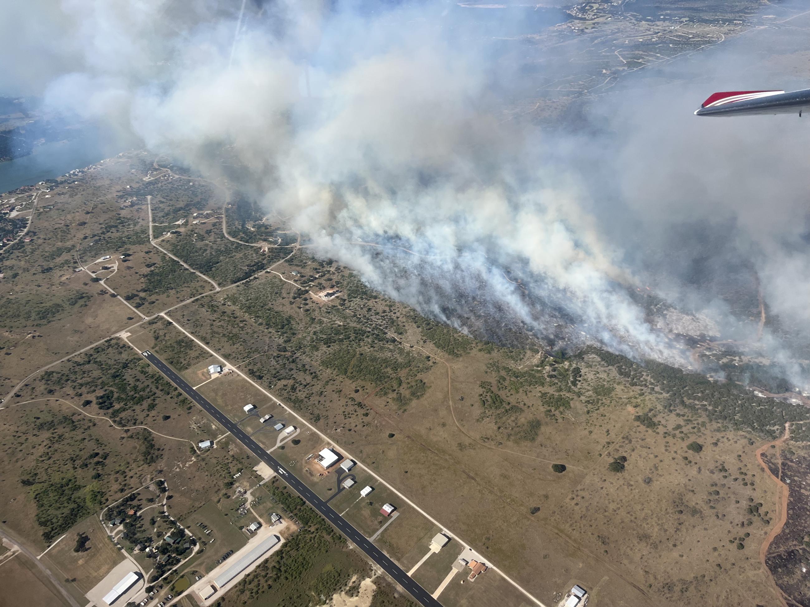 Thick black and gray smoke moving to the left of the photo as fire burns rapidly near a runway