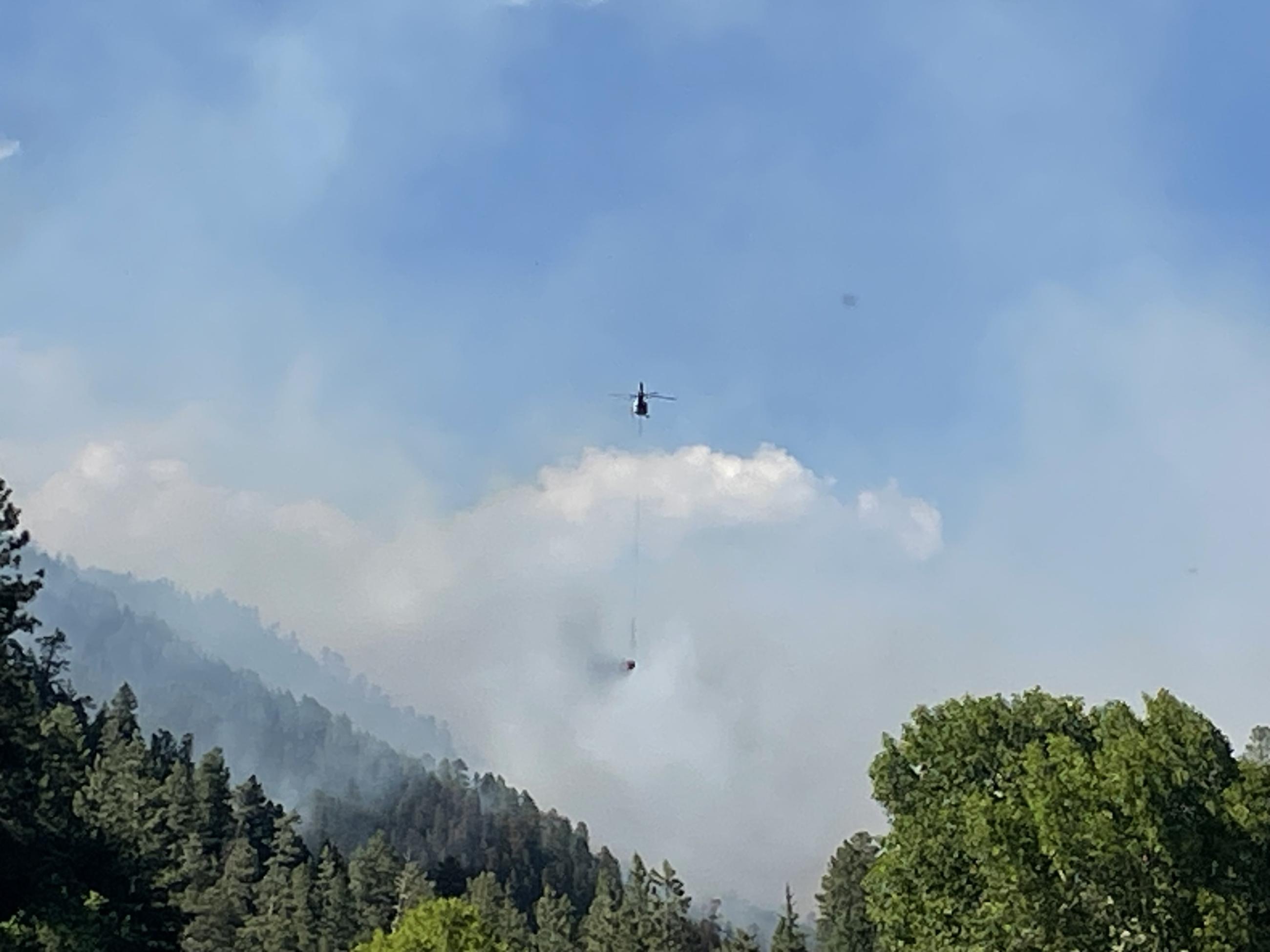 Light helicopter with bucket below and smoke behind