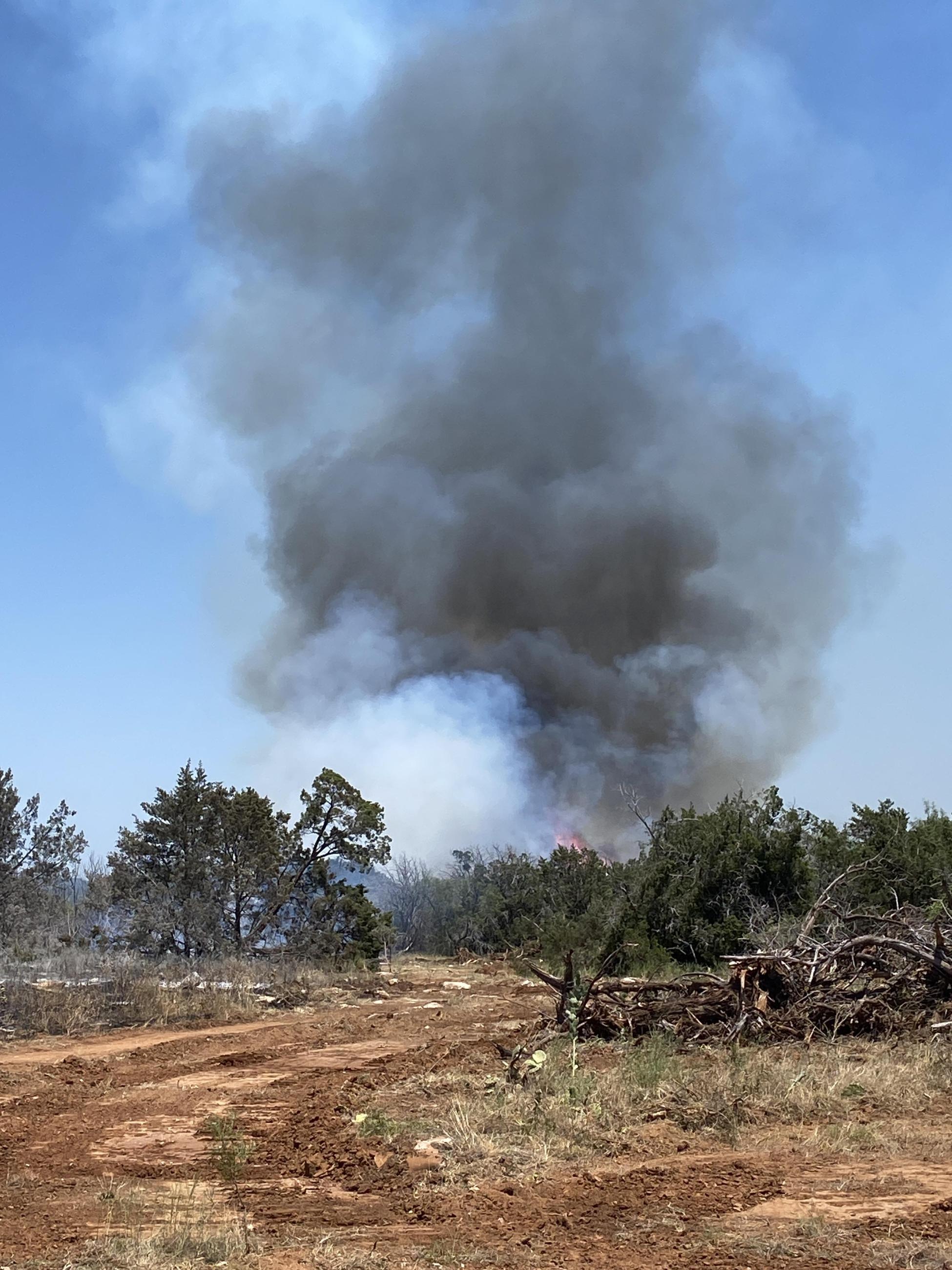 A dark smoke plume rising in the background over a dozer line of bare dirt with grass and brush on the right hand side. The left hand side has already burned and is black and smoldering.