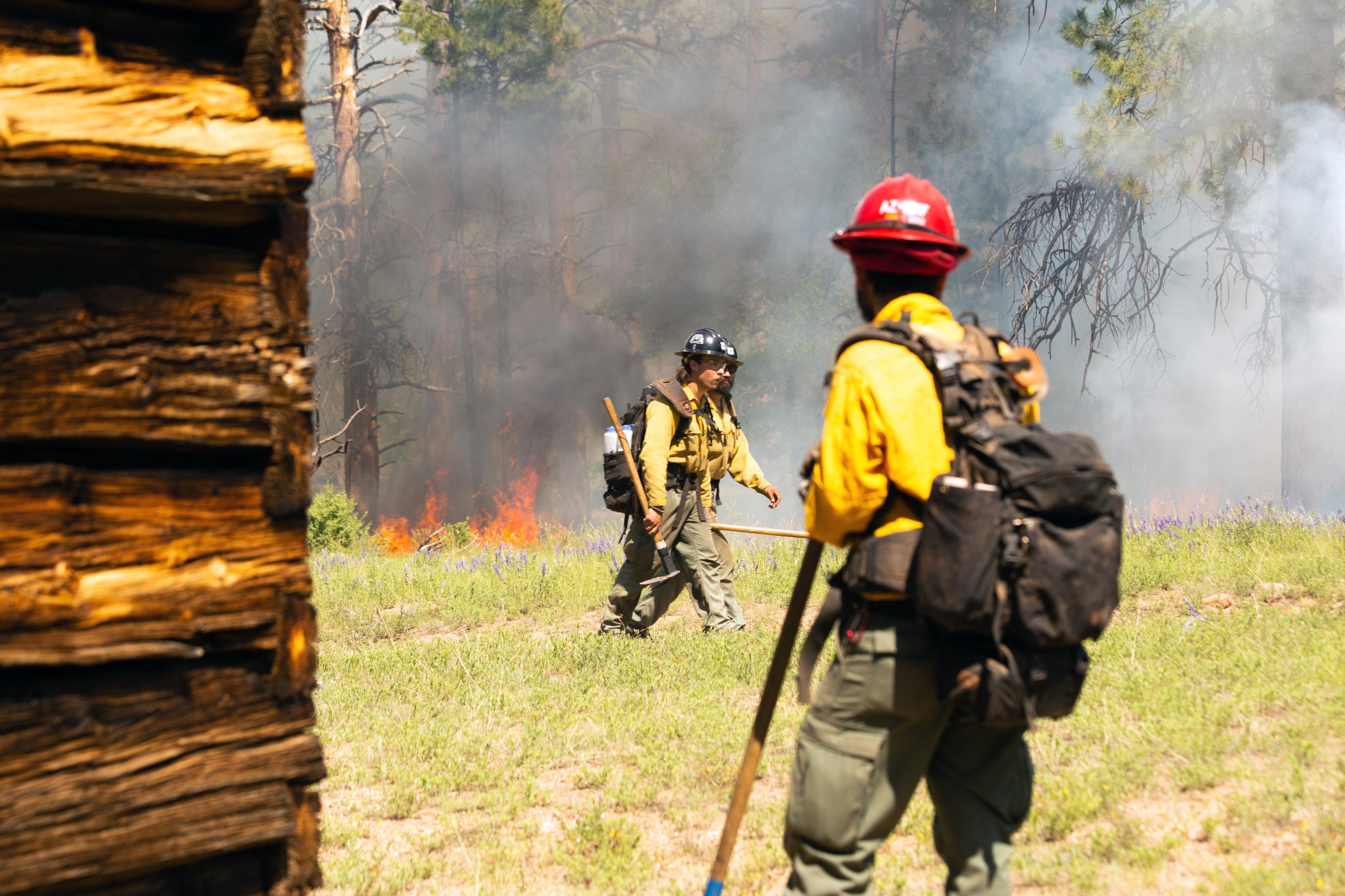 A firefighter wearing a fire pack stands next to a cabin as firefighters walk by and smoke lingers in the background.