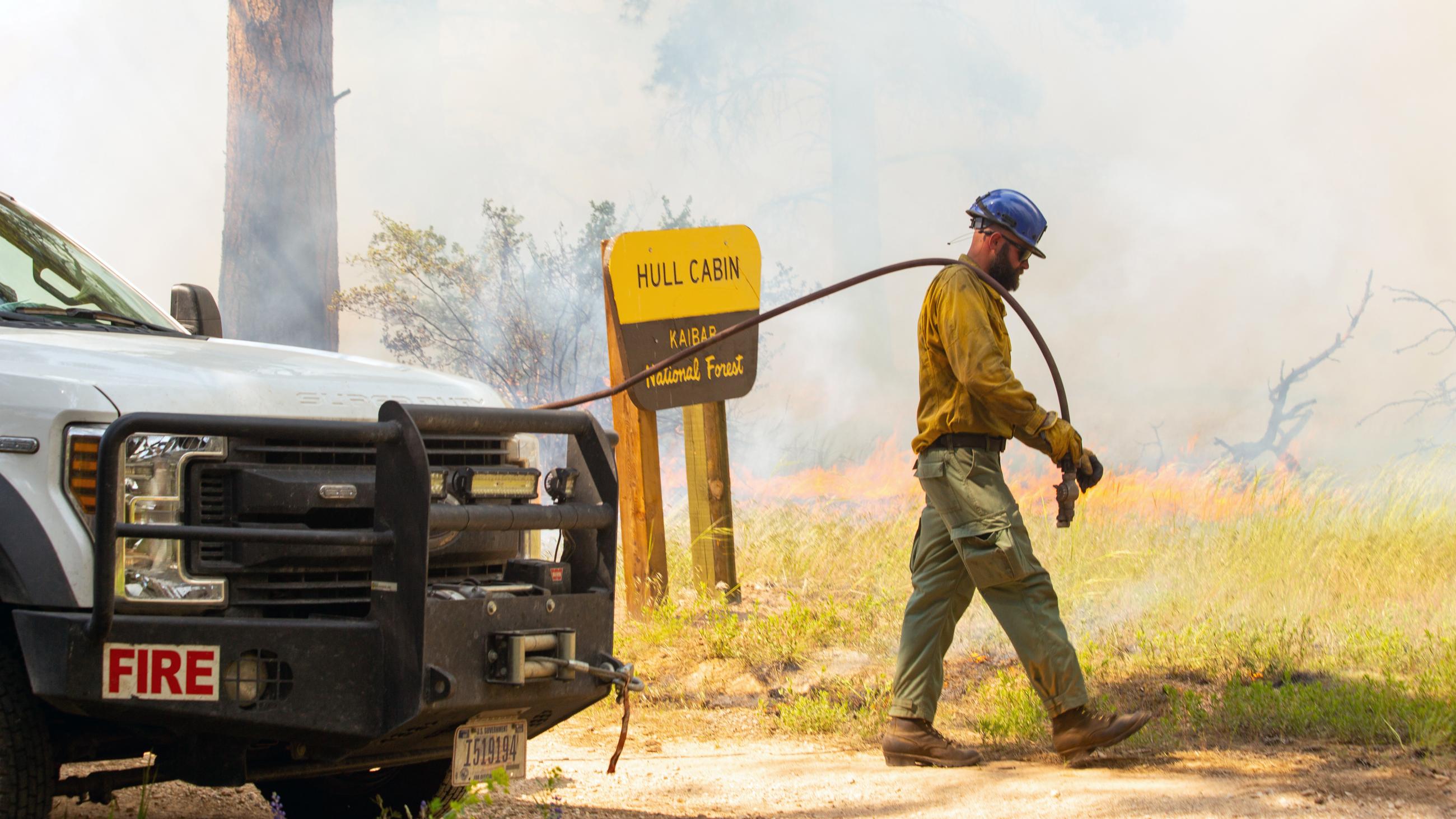 A firefighter wearing a yellow Nomex shirt carries a hose from a fire engine to the fire line while smoke drifts in front of a Hull Cabin sign in the background