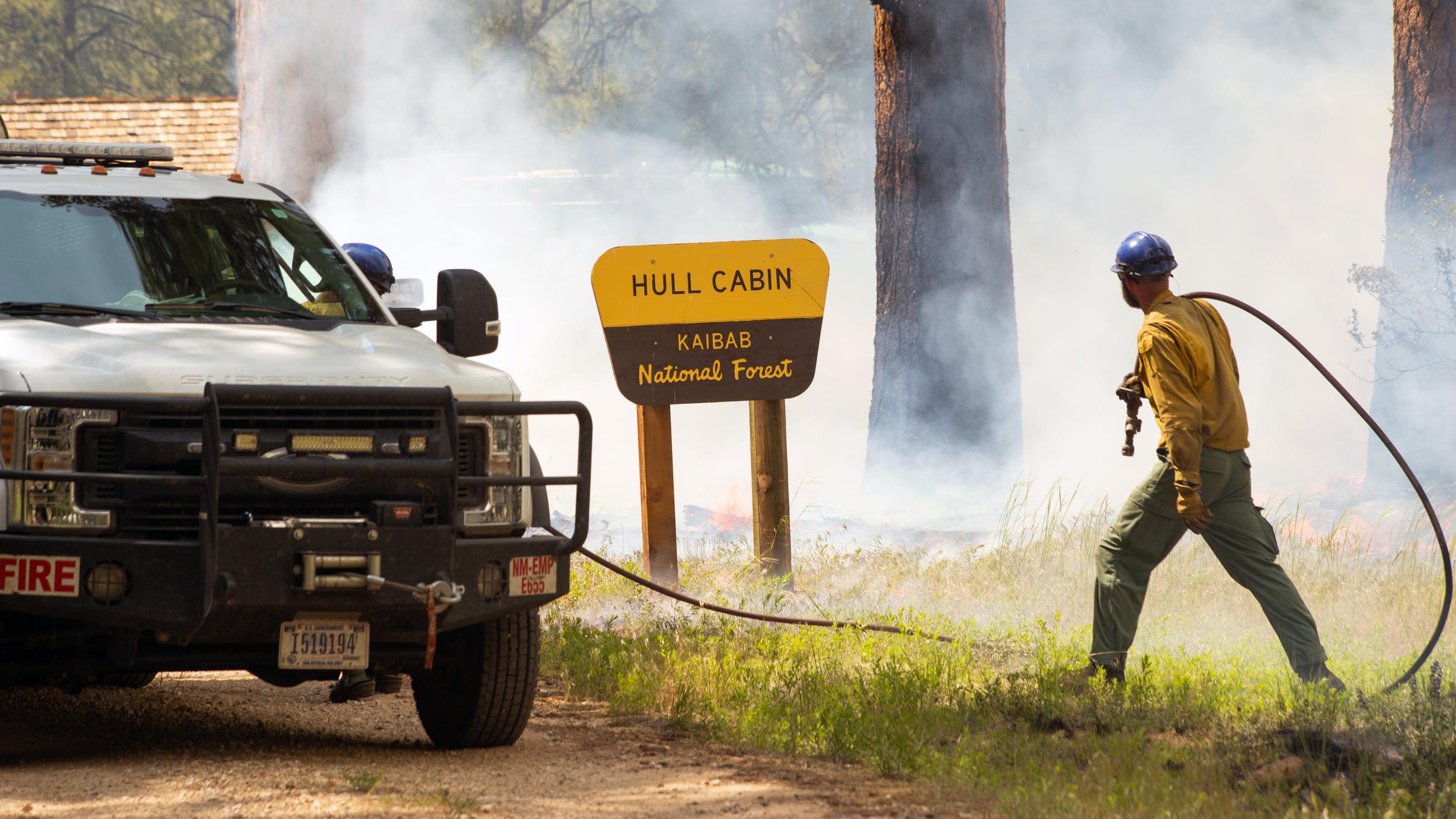 A wildland firefighter carries a hose to an engine in front of the Hull Cabin sign while smoke billows in the background