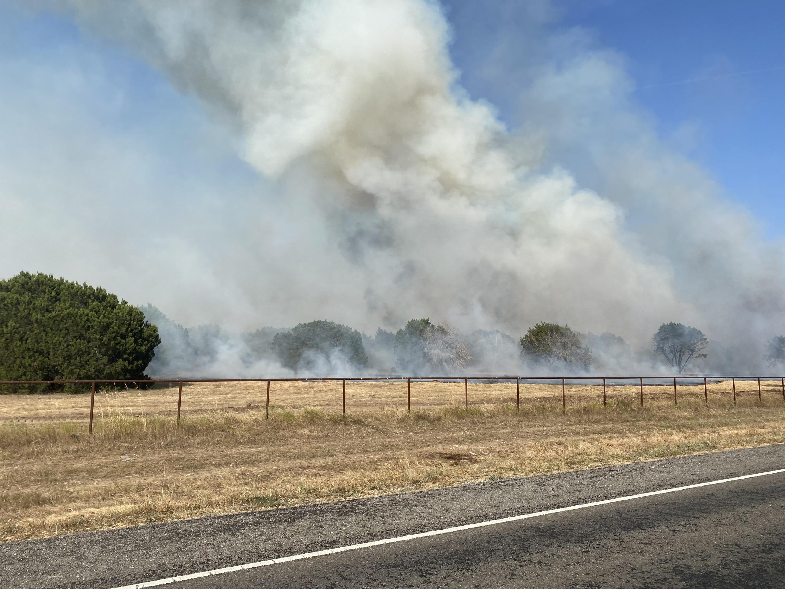 Grey-ish white smoke rises from green juniper in the back ground. Brown grass, a fenceline and a paved public road in the forground